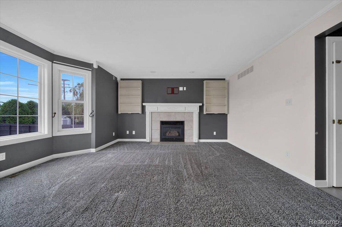 Unfurnished living room with a fireplace, built-in shelves, gray carpet, and large windows.