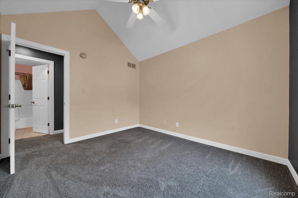 Empty bedroom with gray carpet, beige walls, vaulted ceiling, and an open doorway to the bathroom.