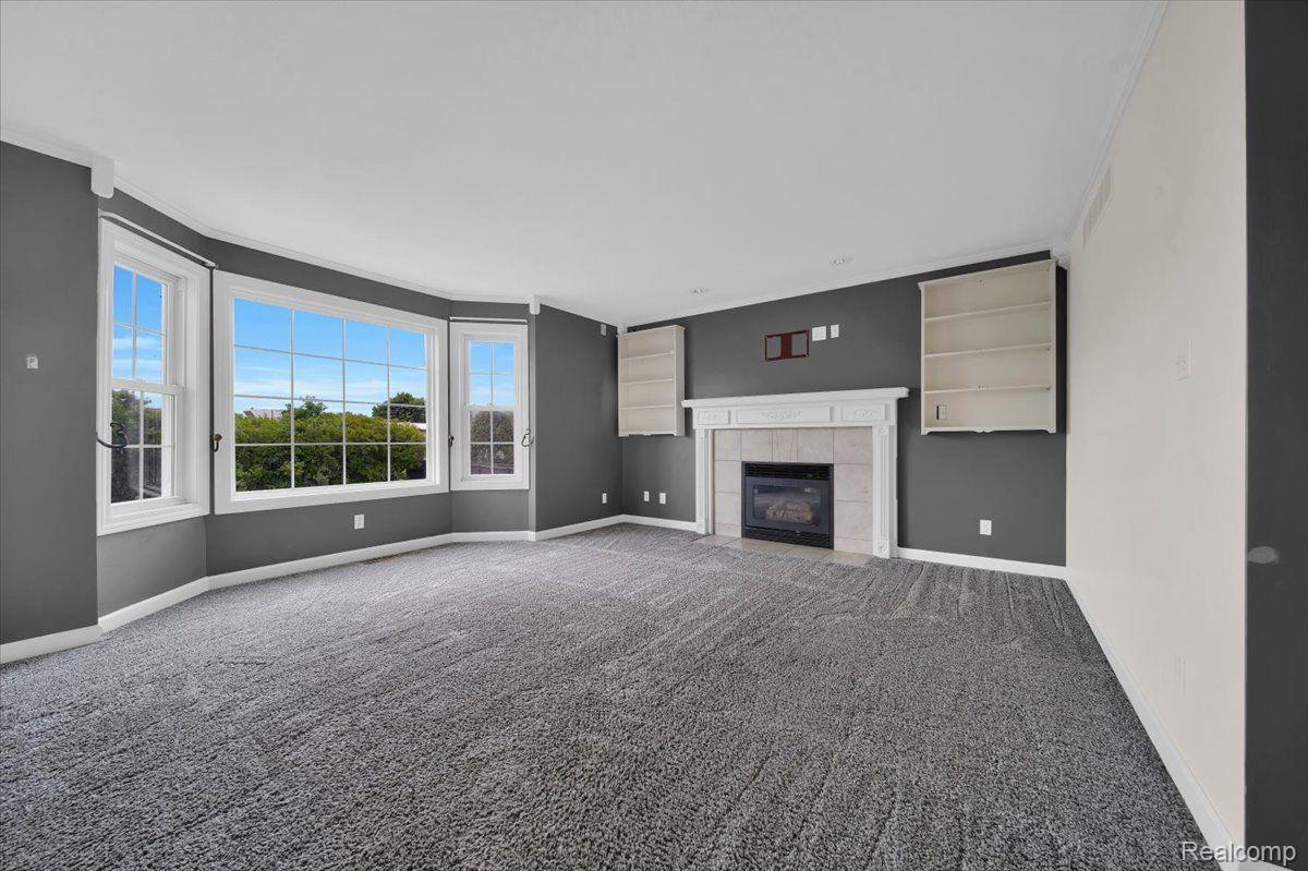 Empty living room with a white fireplace, built-in shelves, gray carpet, and large windows.