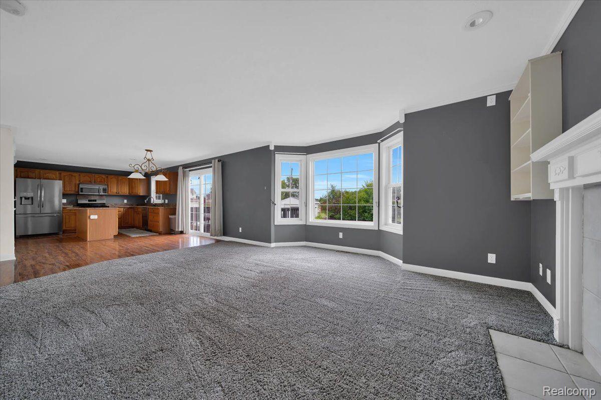 Empty living room with gray carpet, bay window, and open kitchen with wood cabinets.