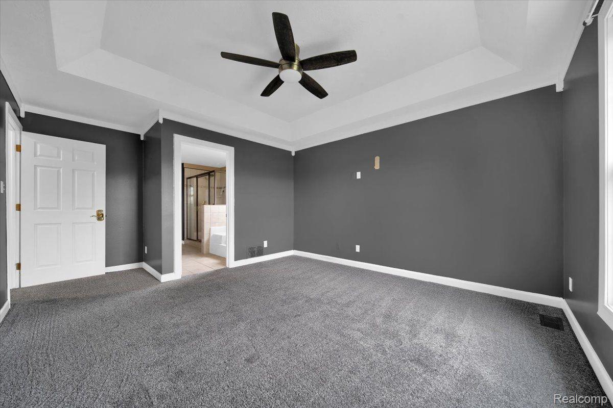 Empty bedroom with gray walls, carpet, tray ceiling, ceiling fan, and a doorway to the bathroom.