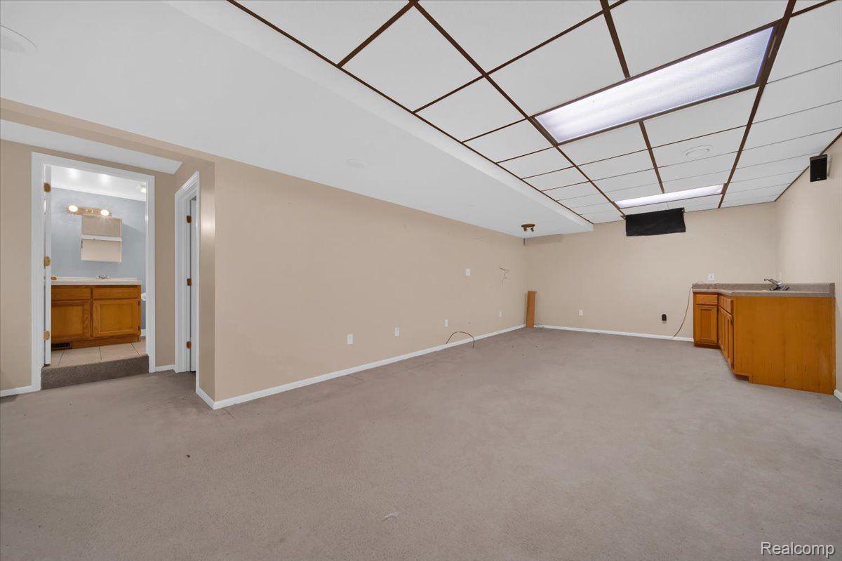 Empty finished basement with beige carpet, a suspended ceiling, and a wood kitchenette.