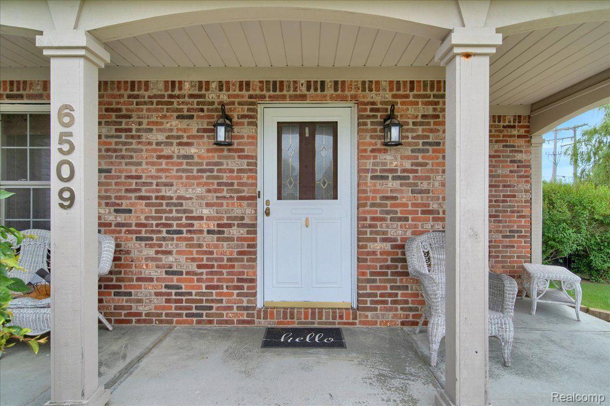 Brick home’s covered front porch with a white door, wicker seating, and house number 6509.