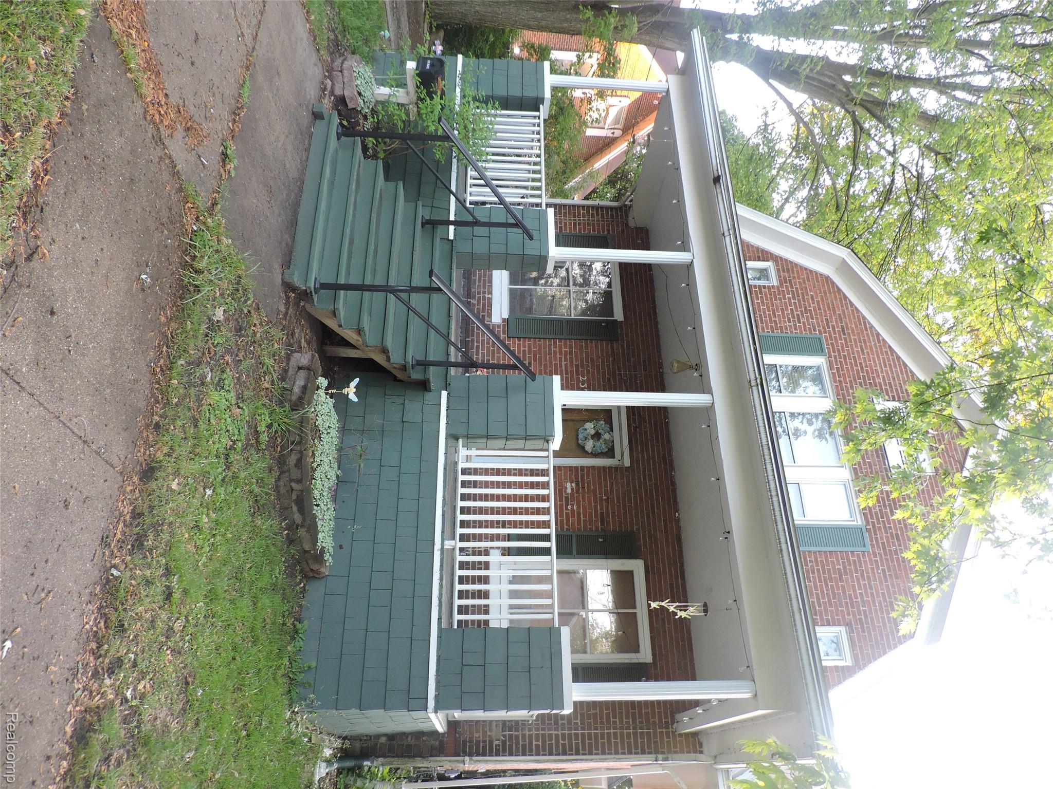 Brick house with a deep covered porch, white railings, and broad green steps beneath mature trees.