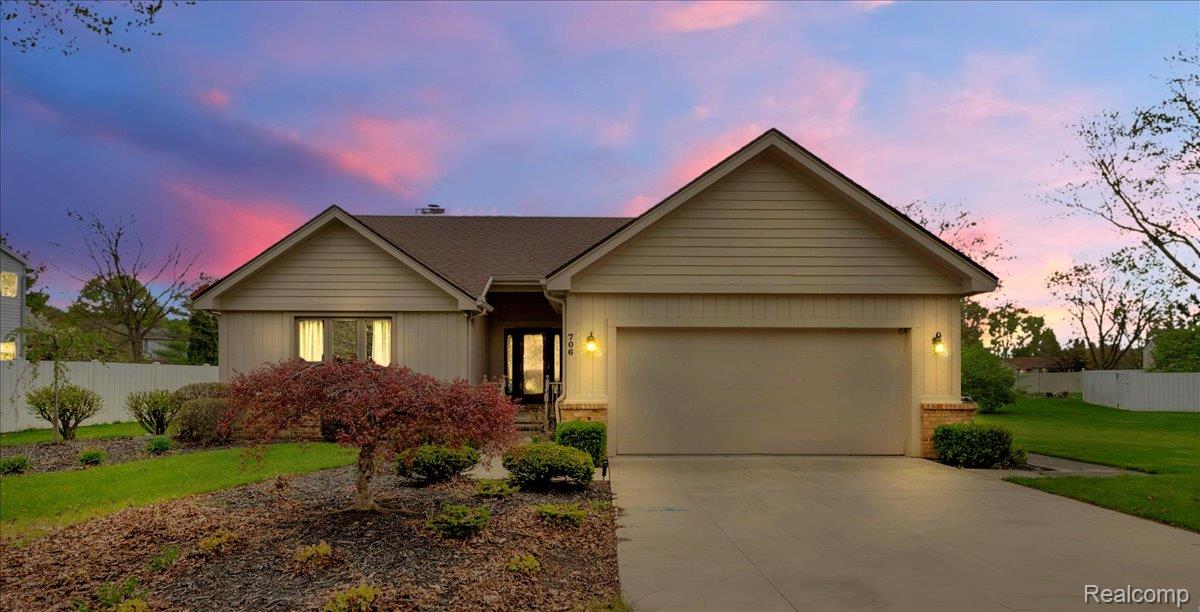 Single-story beige house with a two-car garage and landscaped front yard at dusk.
