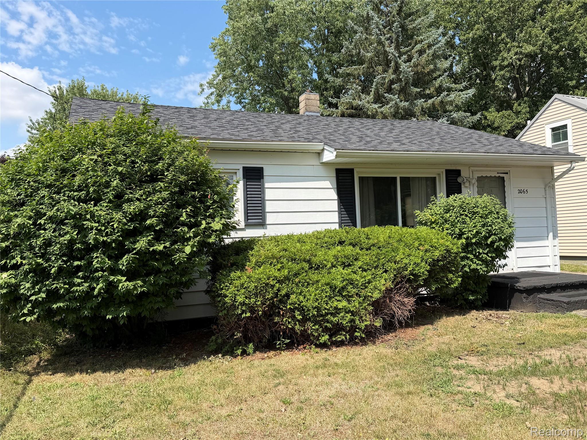 White single-story house with black shutters, a dark roof, and large shrubs across the front.