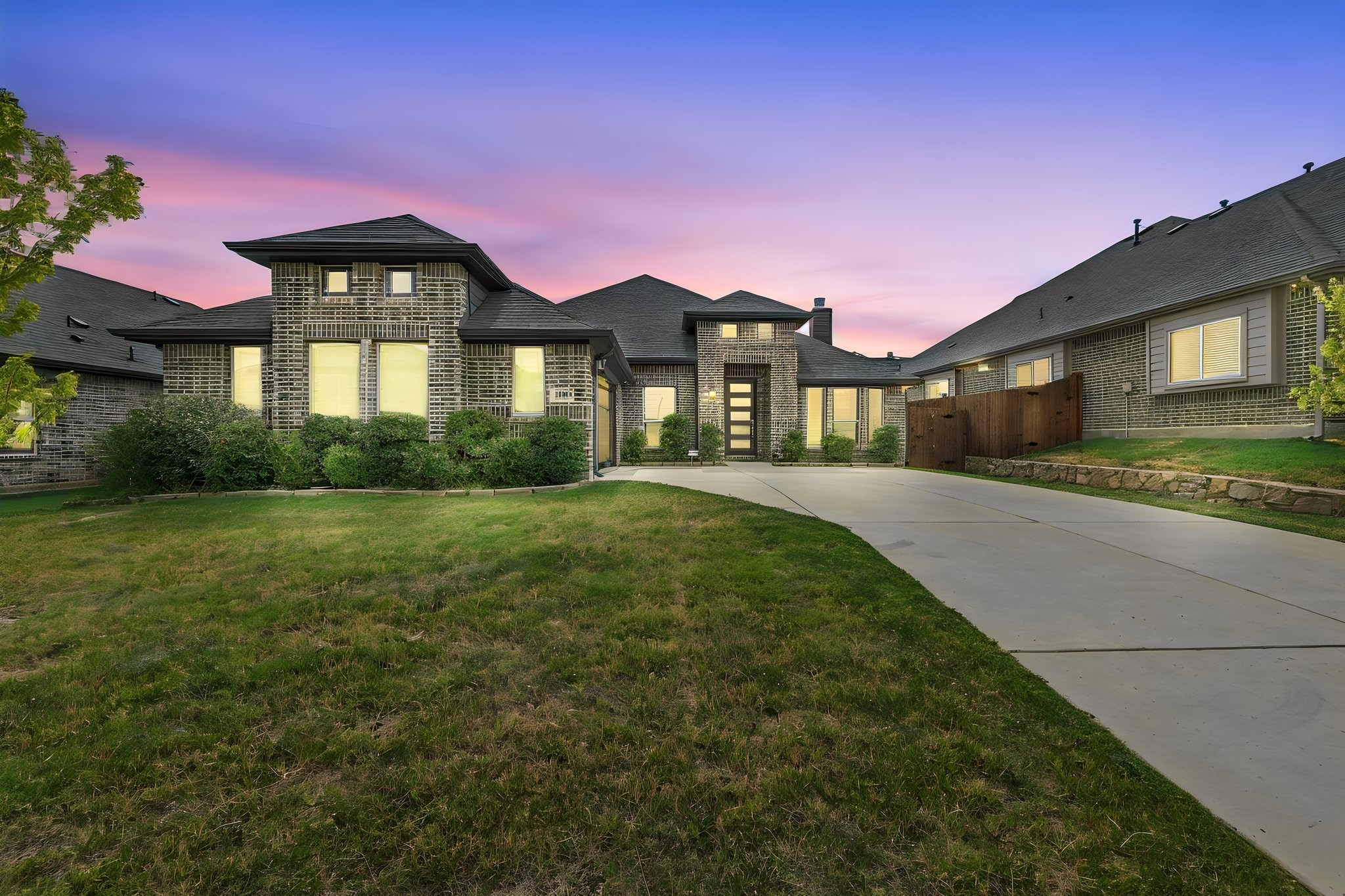 Large brick home with glowing windows, a broad driveway, and lawn beneath a pink dusk sky.