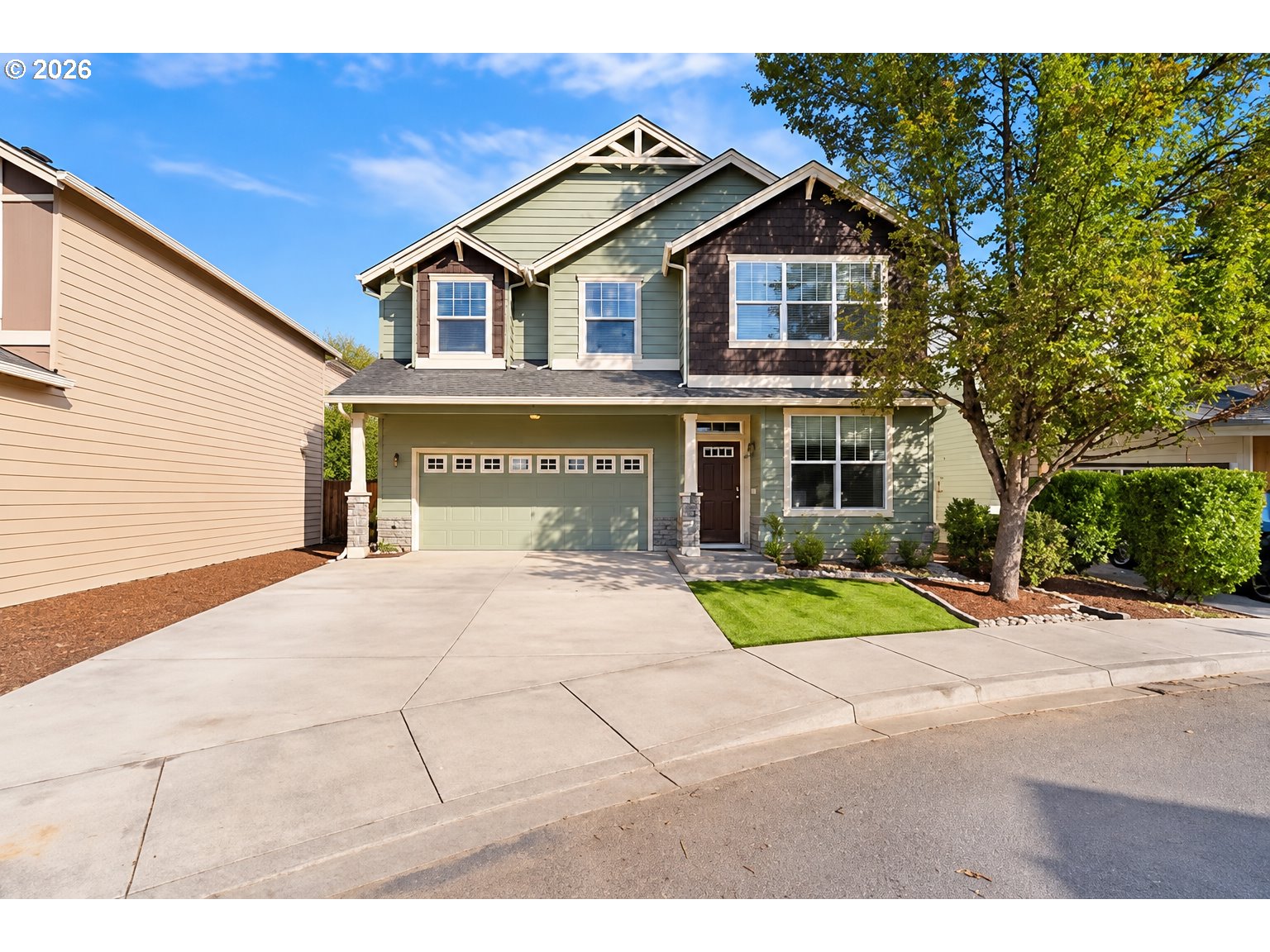 Two-story green house with a double garage, broad driveway, and mature tree in front.