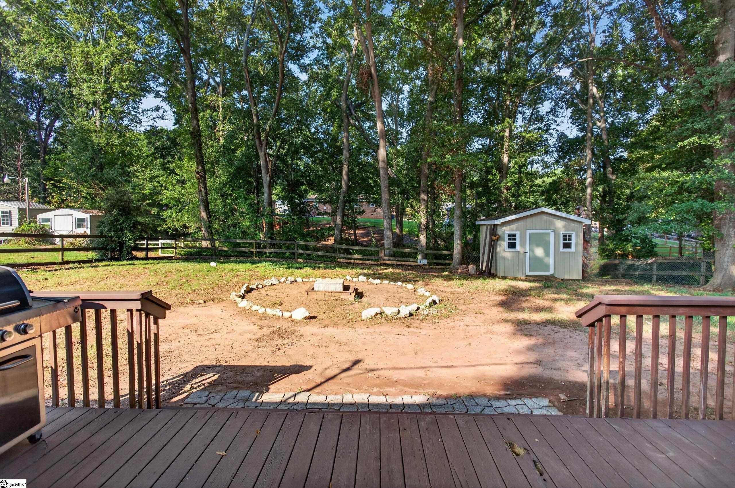Wooden rear deck overlooking a fenced, tree-shaded yard with a shed and stone-ringed fire pit.