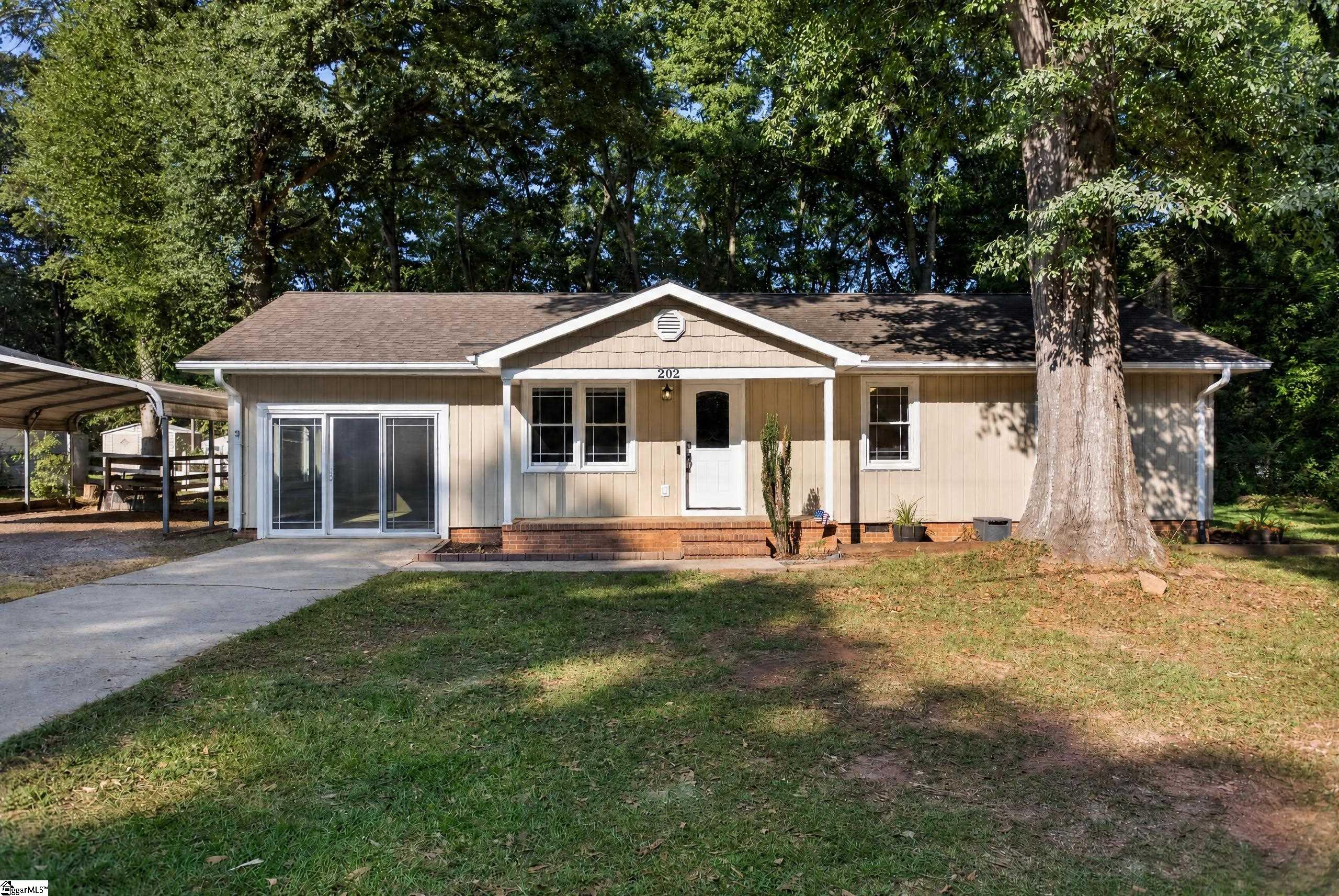 Single-story tan house with sliding glass doors, a covered entry, and a carport beneath mature trees.
