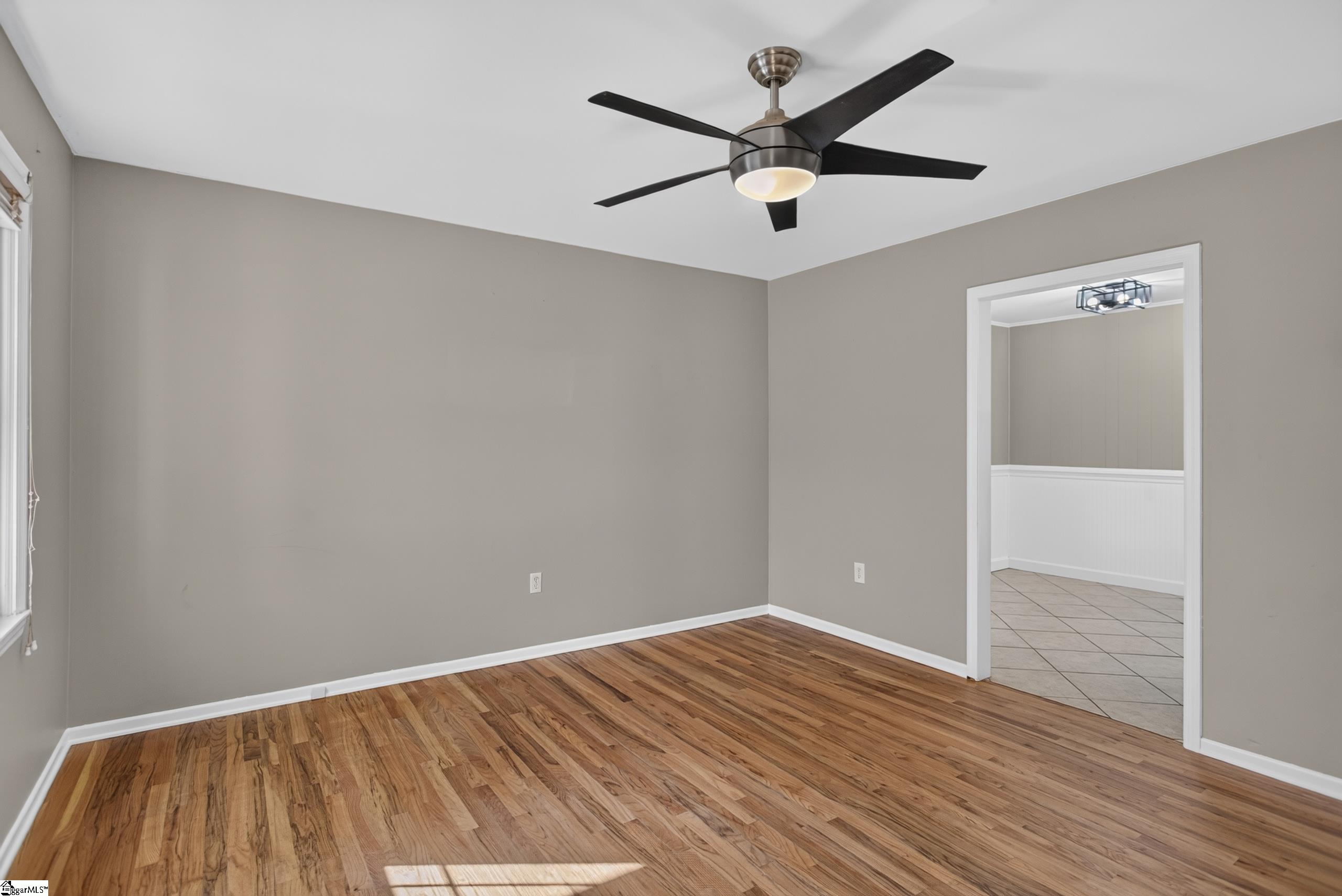 Vacant room with hardwood floors and a ceiling fan, opening into a tiled adjoining room.