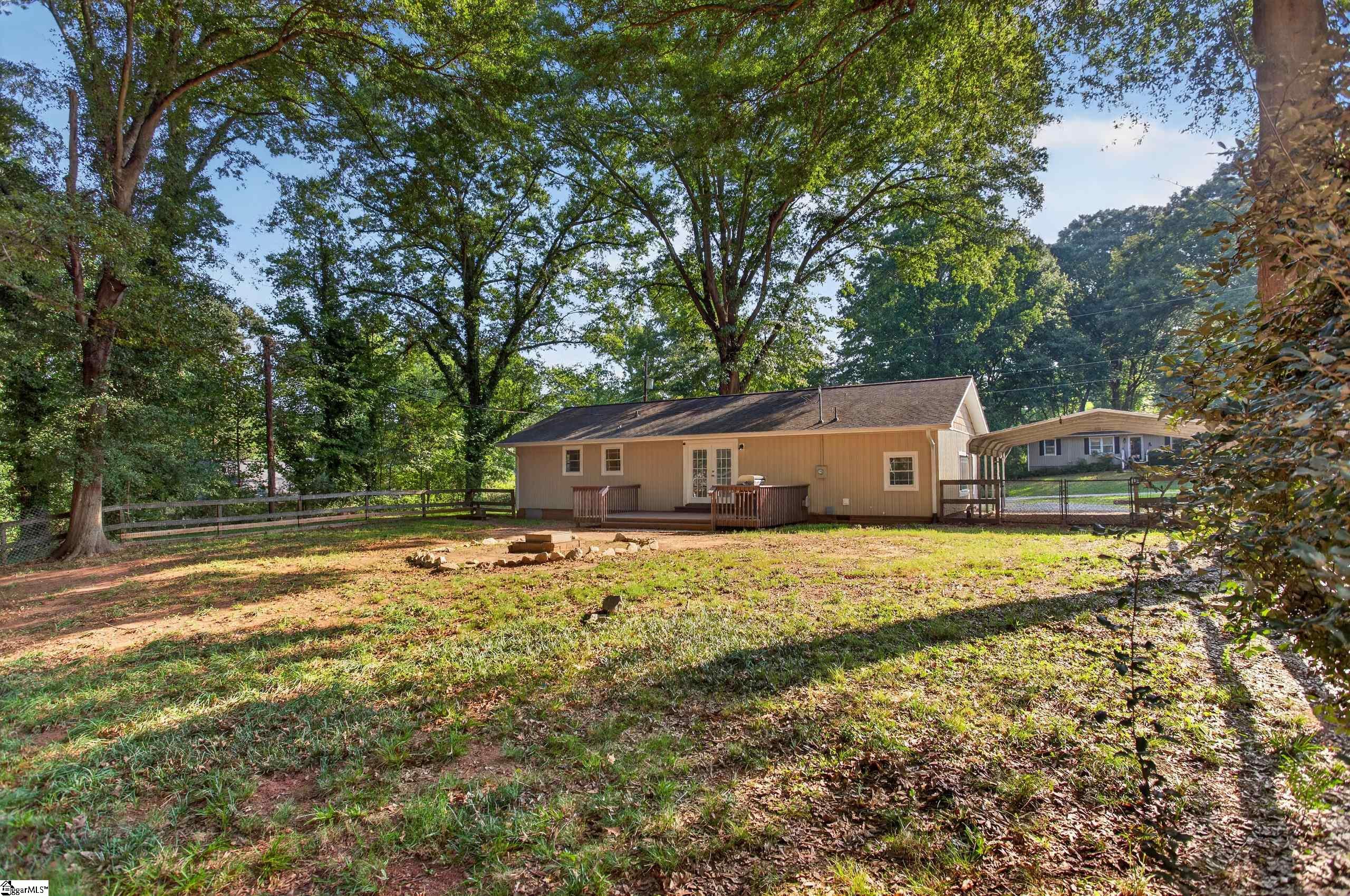 Tan single-story home with a small deck and fire pit in a wooded backyard.