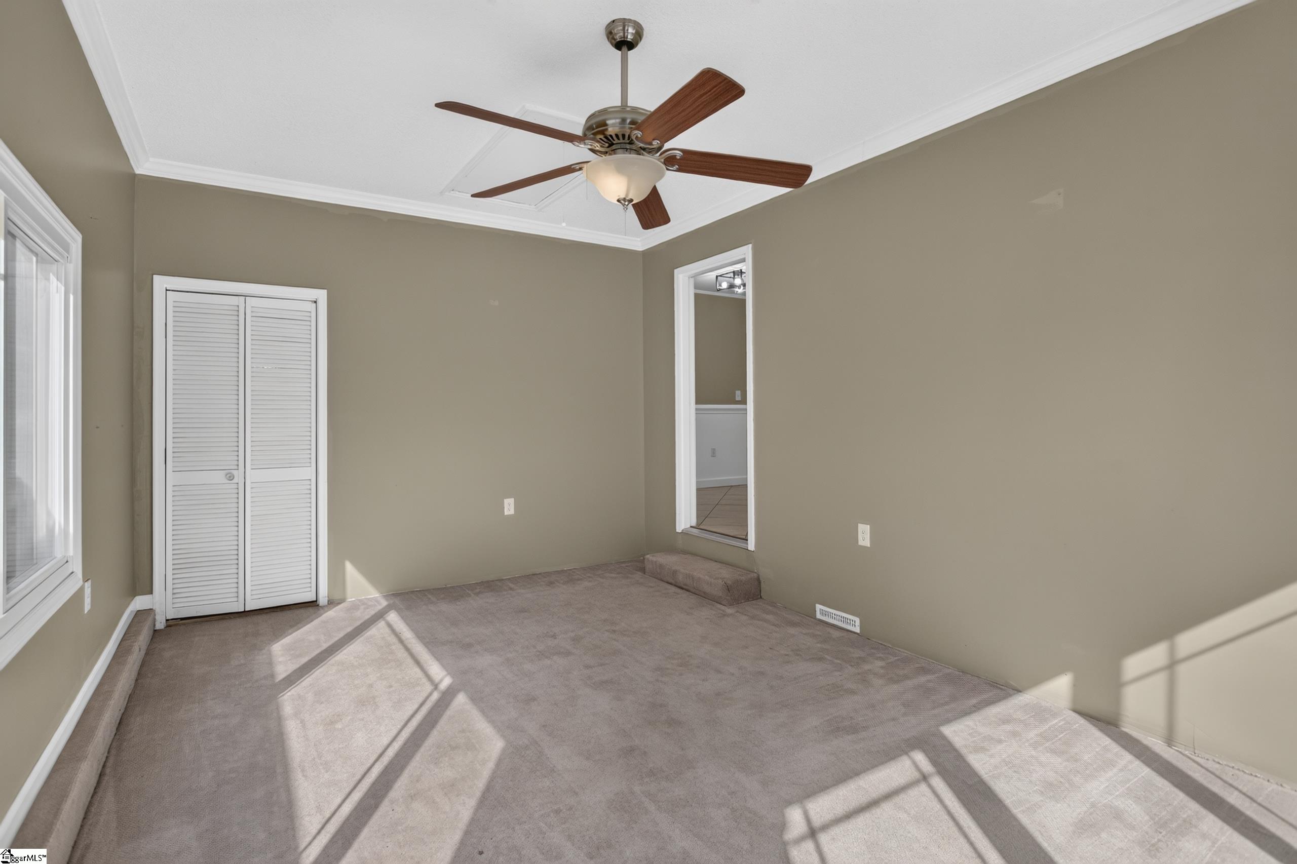Empty carpeted bedroom with a ceiling fan, louvered closet, and bright sunlight on the floor.