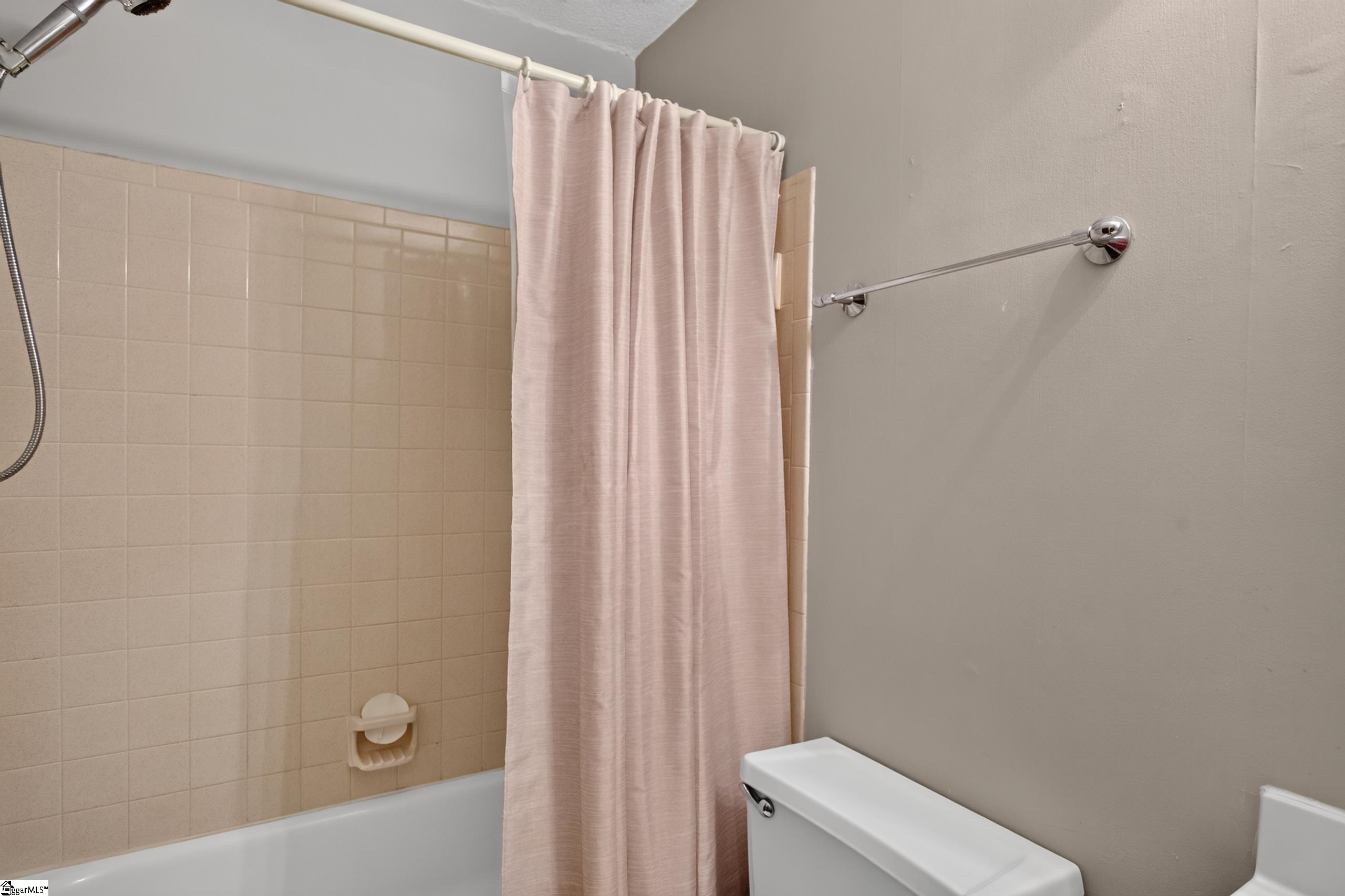 Bathroom with a beige tiled tub and shower, pale pink curtain, and white toilet.