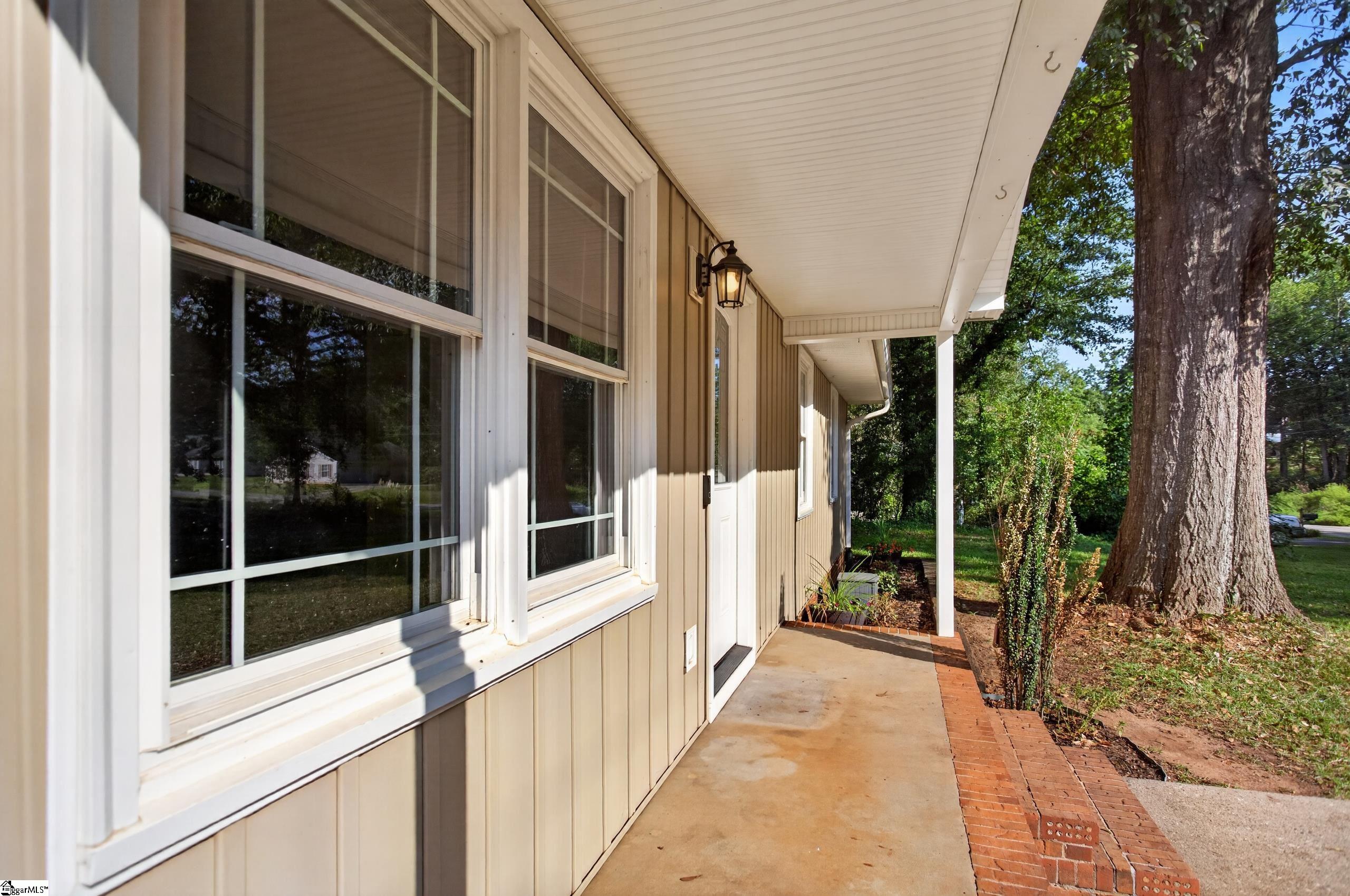 Covered porch with white-trimmed windows and brick edging beside a large mature tree.