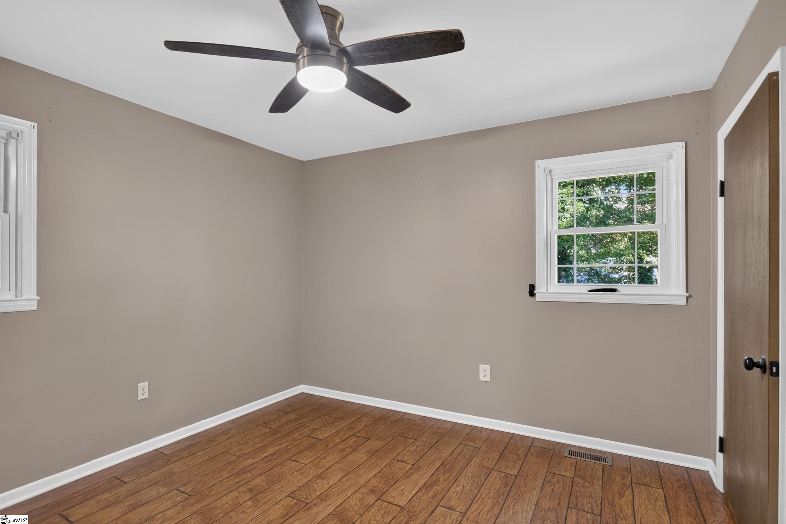 Empty bedroom with wood-look flooring, a dark ceiling fan, and white-trimmed windows.