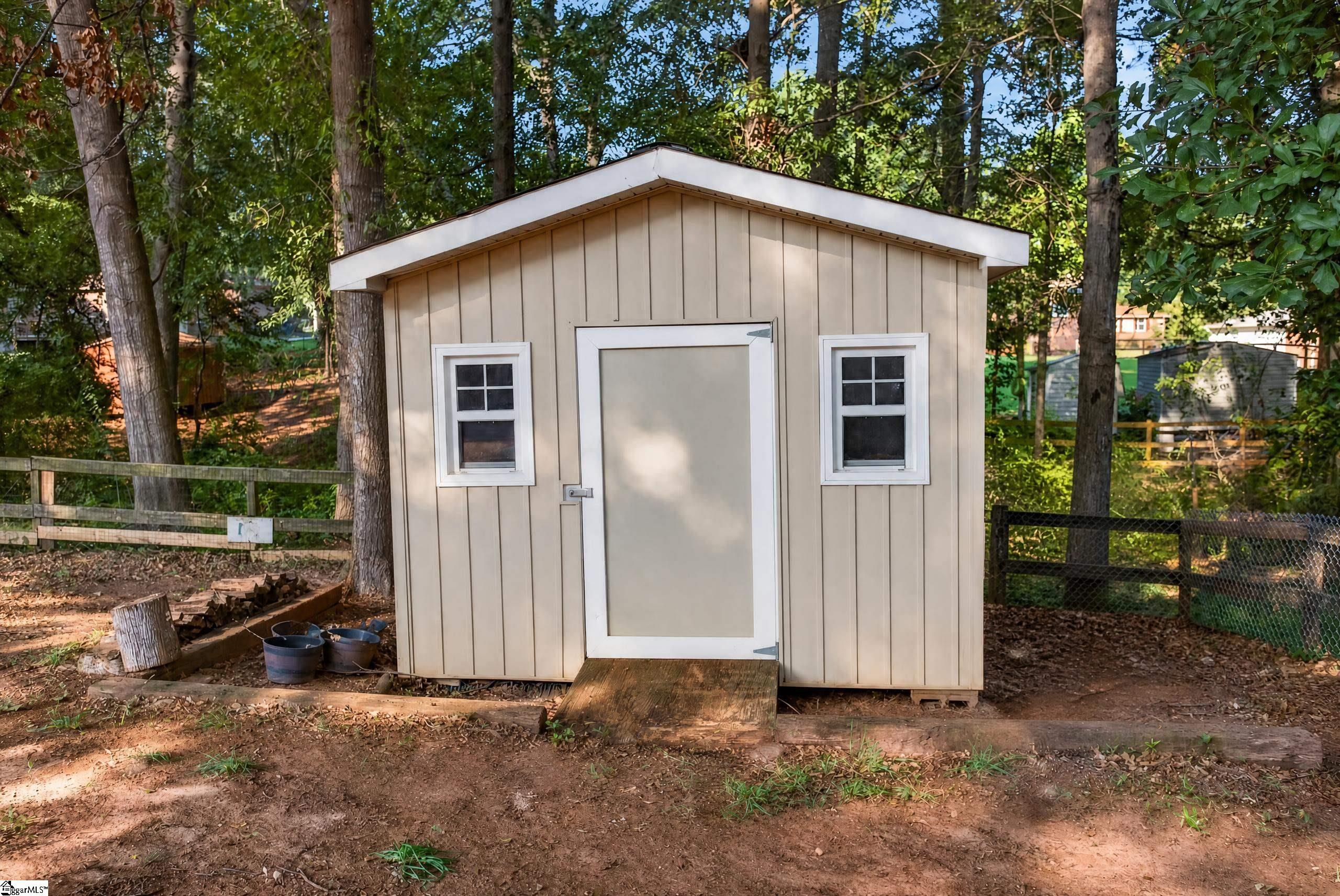 Beige storage shed with white trim, two windows, and a wooden entrance ramp in a wooded yard.