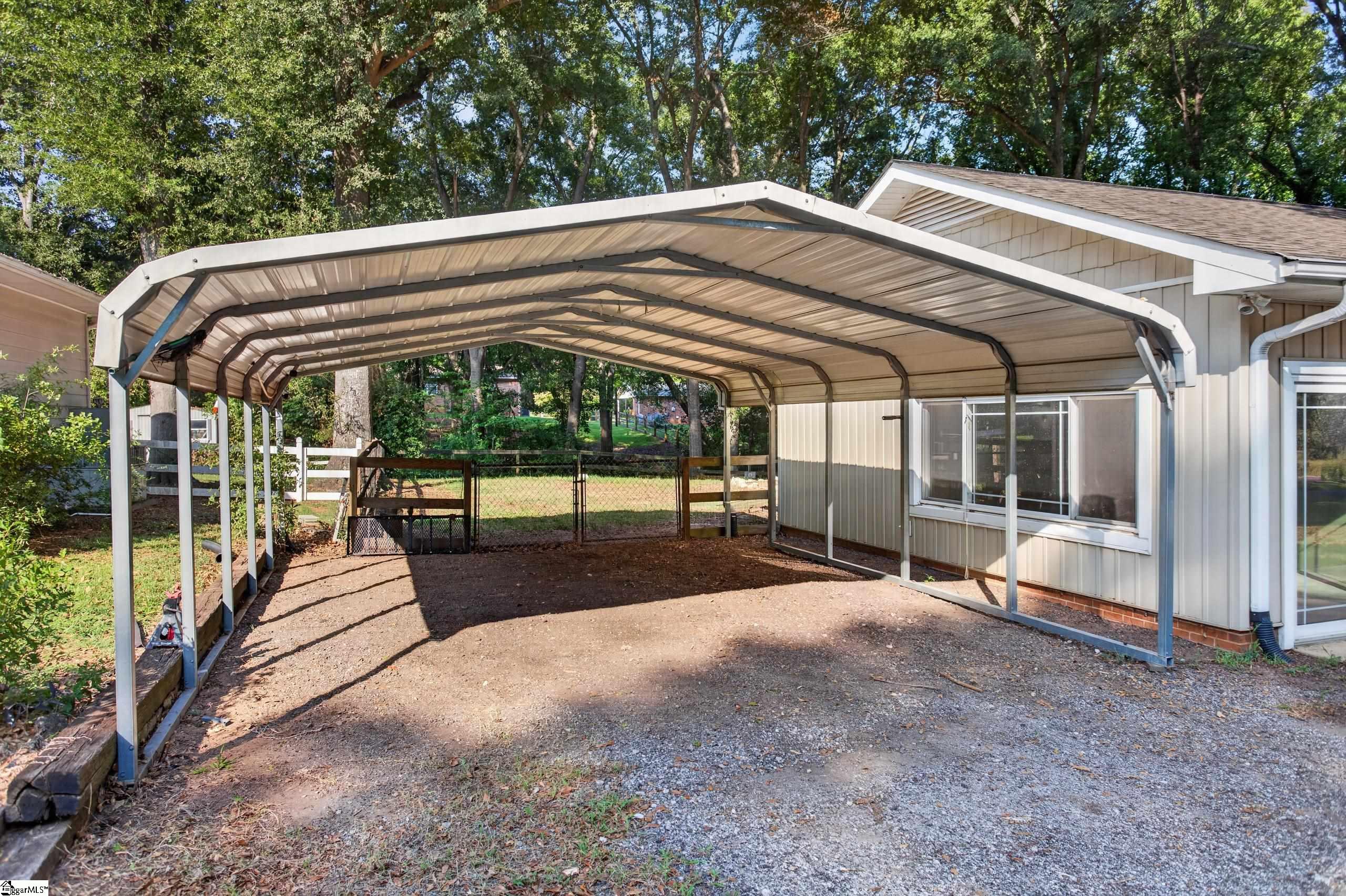 Metal-roofed carport beside a house, with a gravel driveway and fenced, tree-lined yard.