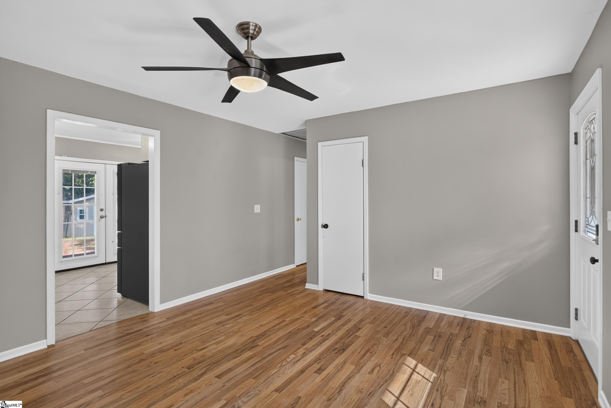 Empty room with hardwood floors, gray walls, a black ceiling fan, and a tiled kitchen doorway.
