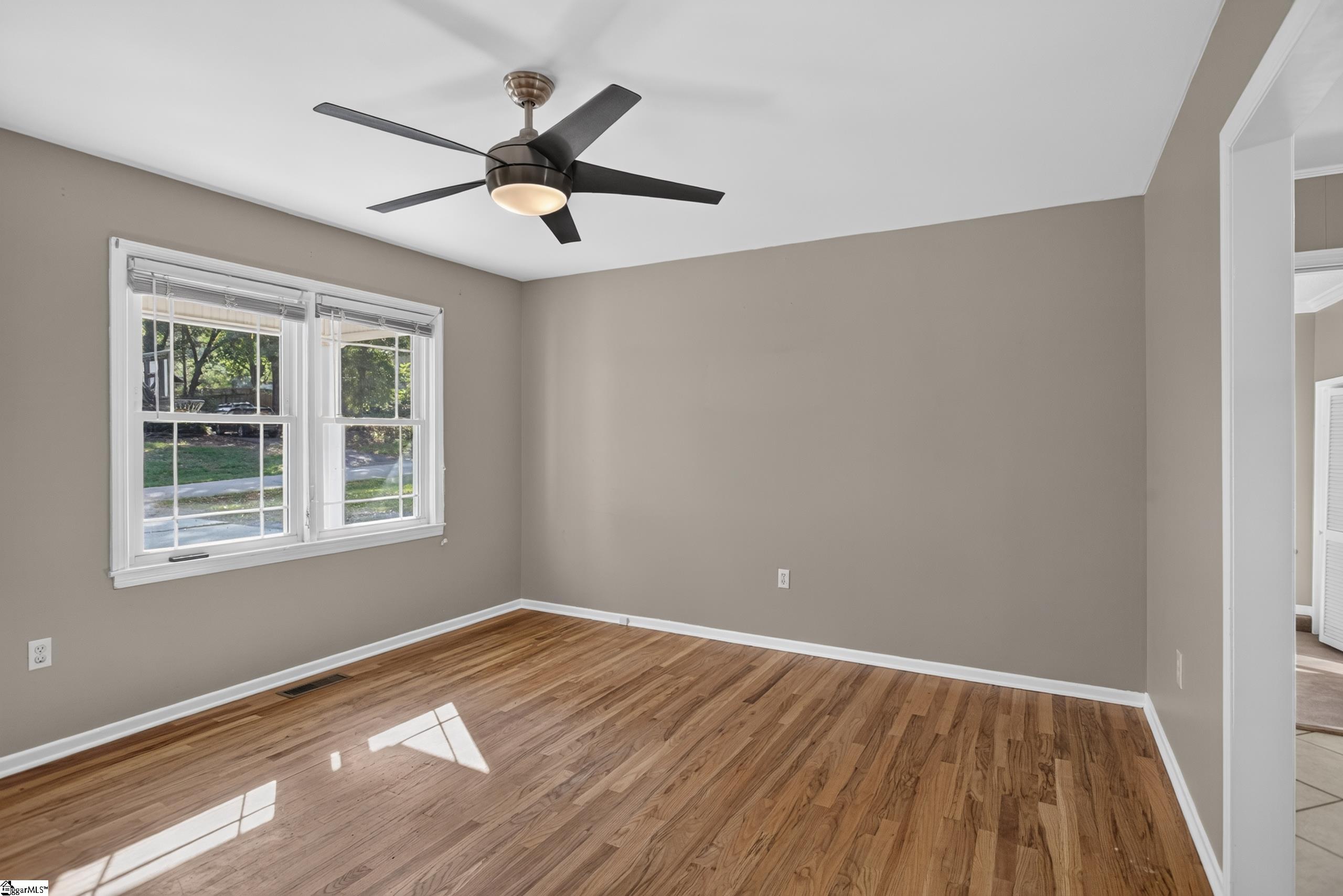 Empty room with hardwood floors, two windows, and a dark ceiling fan.