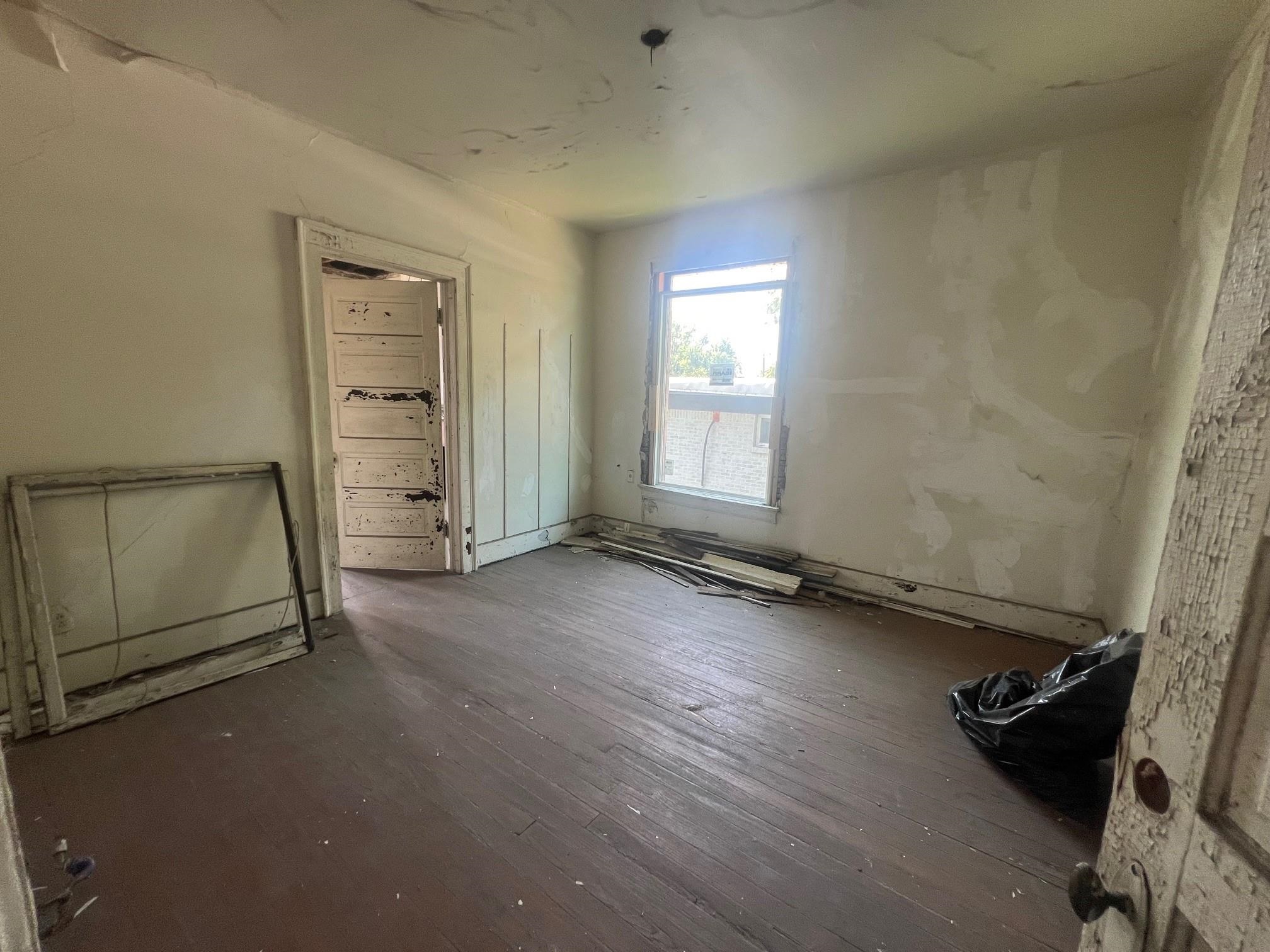 Empty, unfinished room with peeling walls, worn wood floors, and debris beneath a window.
