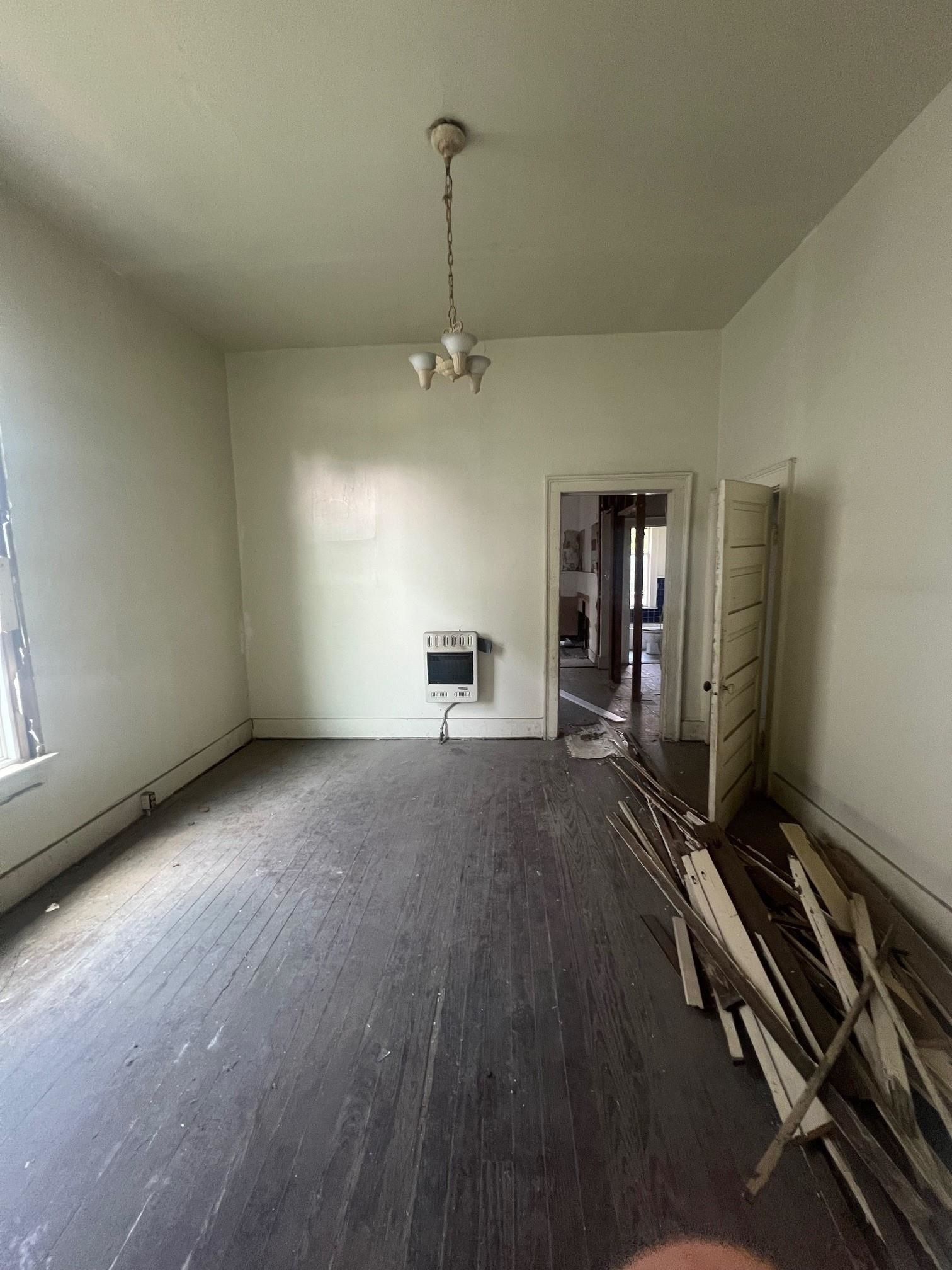 Worn, empty room with a chandelier, dark plank floor, and loose boards piled beside an open doorway.