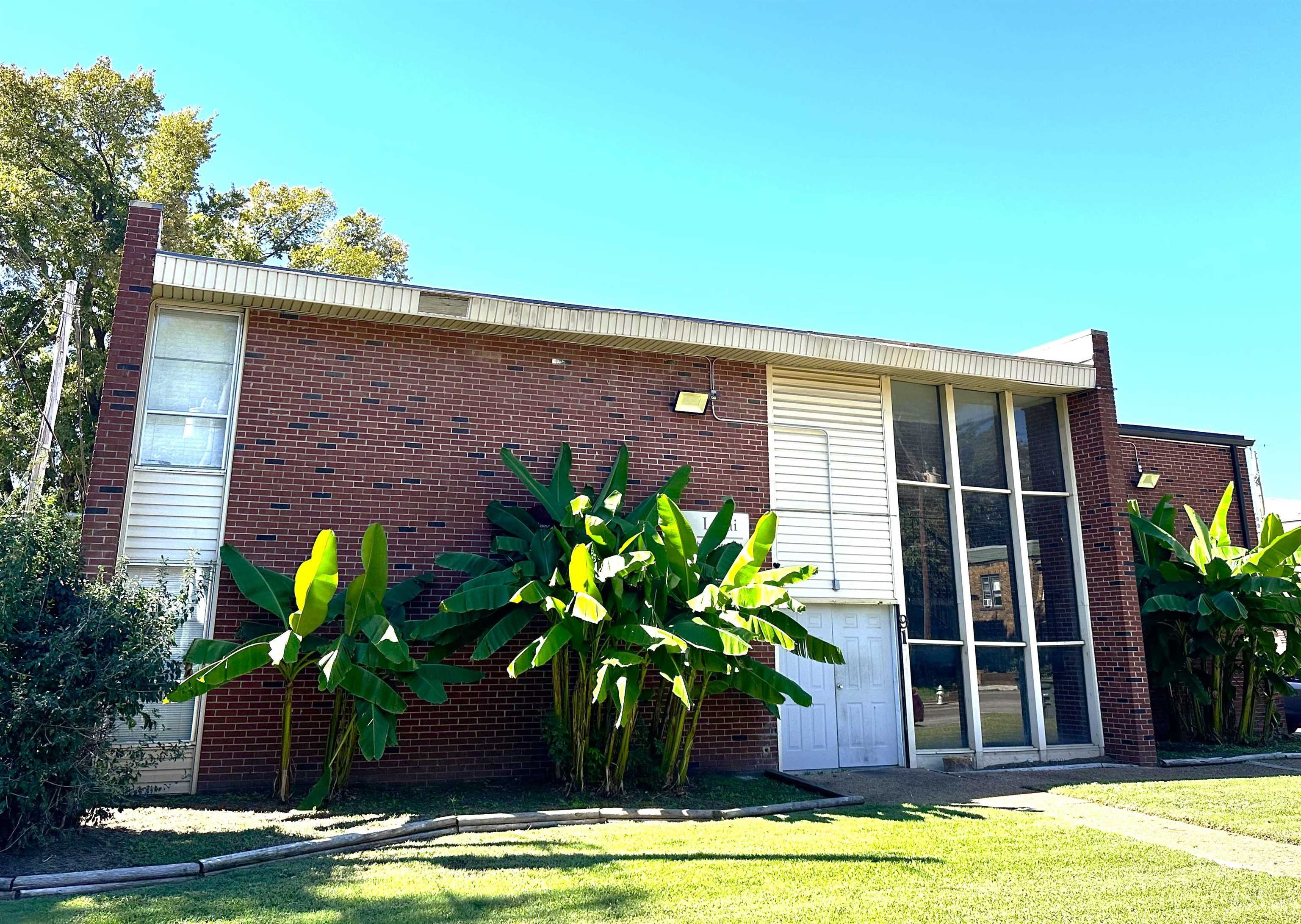 Red-brick residential building with tall windows, tropical plants, and a sunlit lawn.