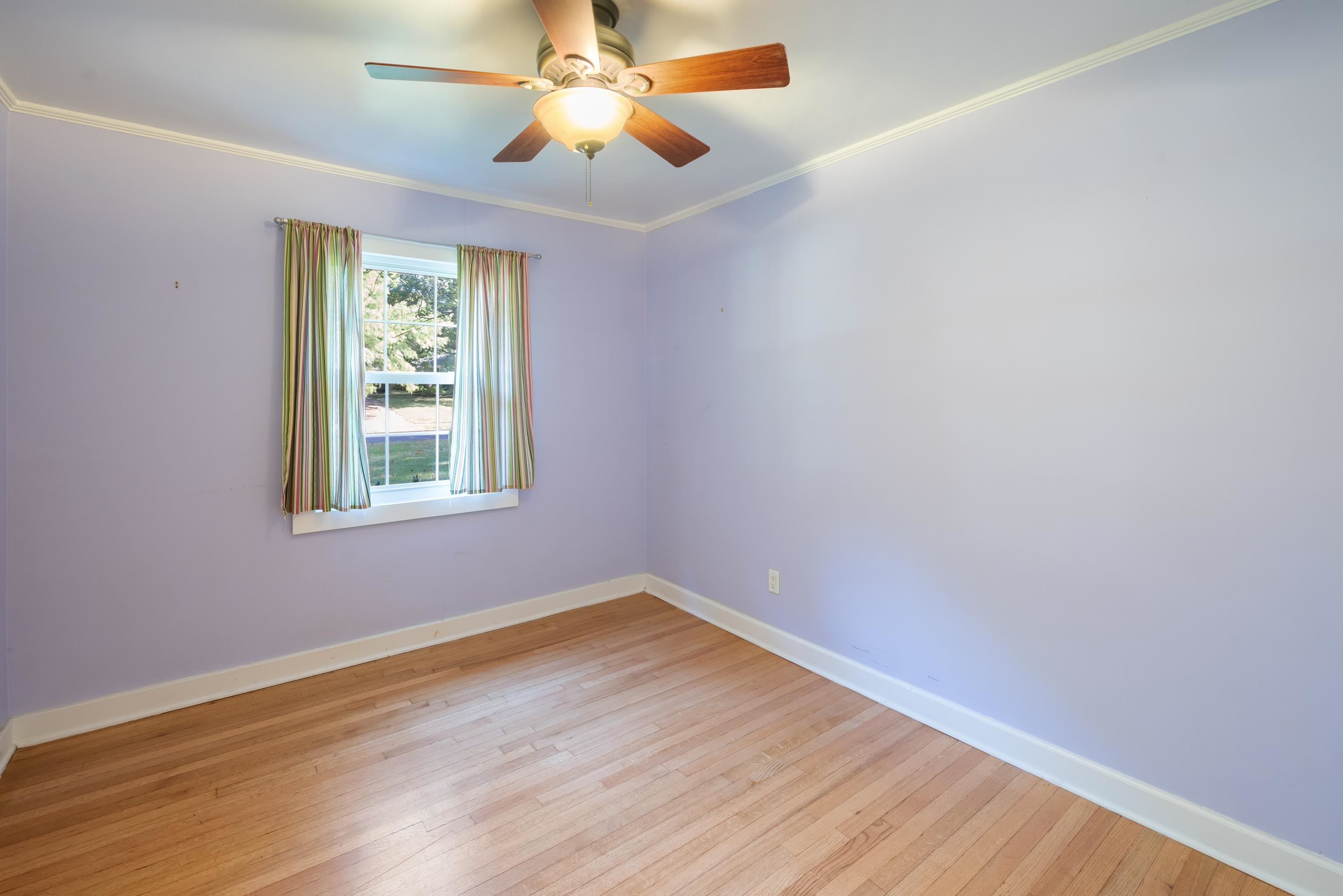 Empty room with lavender walls, hardwood floors, a curtained window, and a ceiling fan.