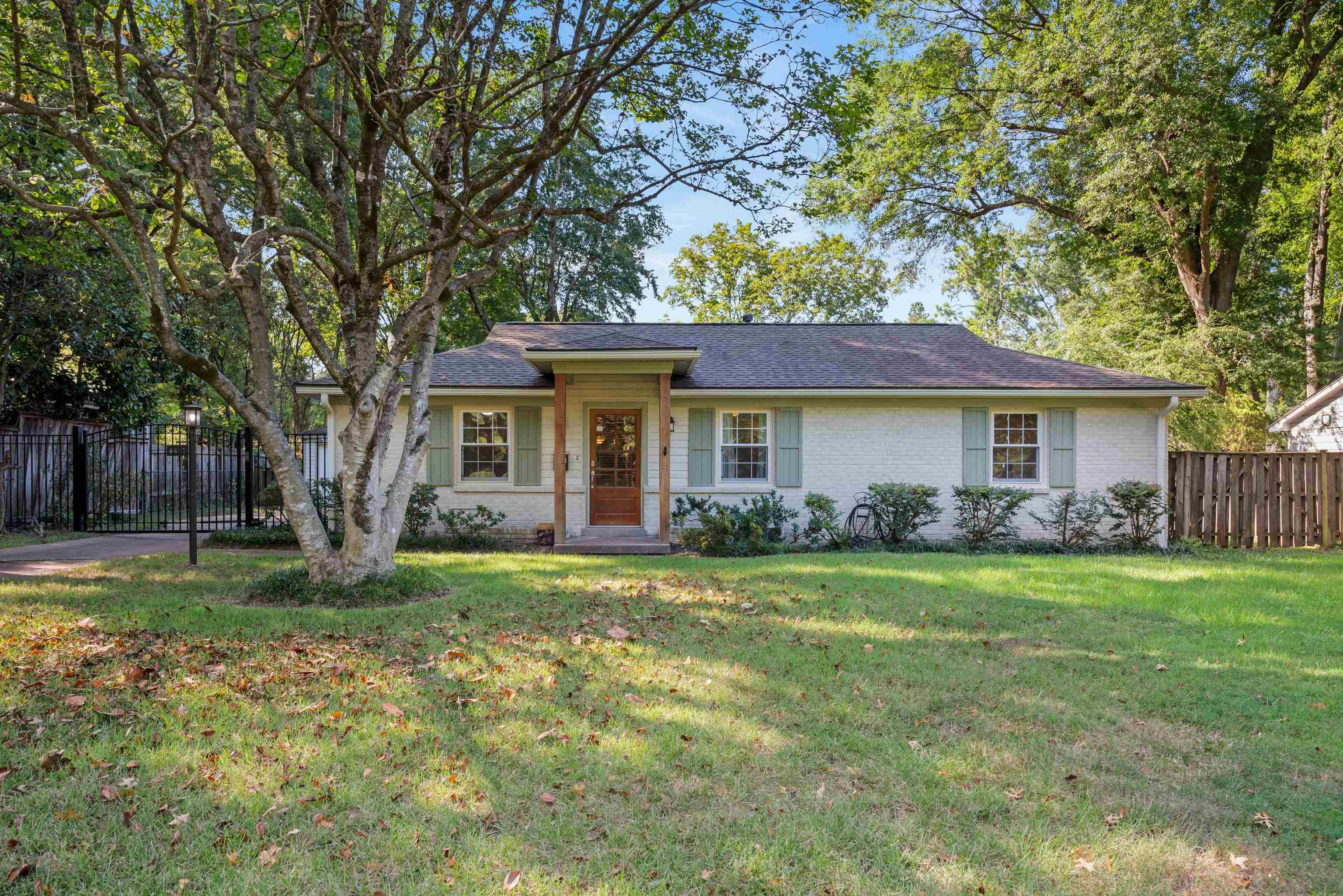 Single-story white brick house with pale green shutters, a covered wood entry, and mature trees across the lawn.