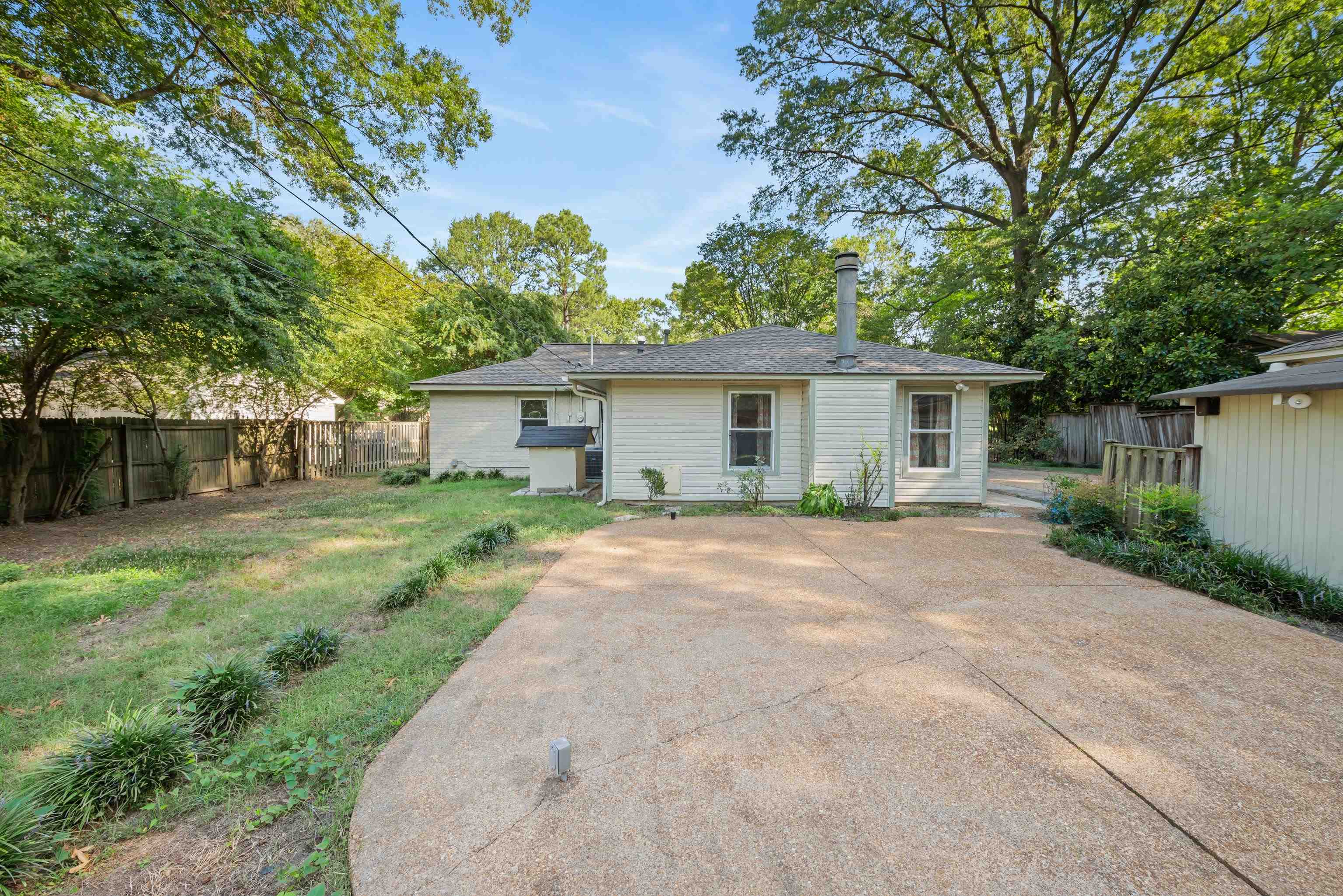 Rear of a single-story house with a wide concrete driveway, mature trees, and fenced yard.