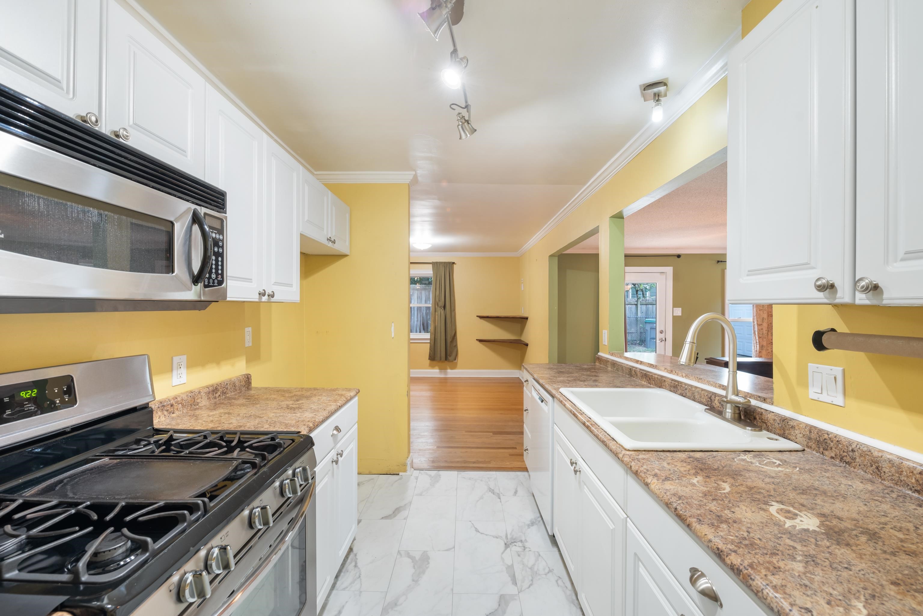 Kitchen with white cabinets, gas range, and patterned counters opening to a hardwood-floored room.