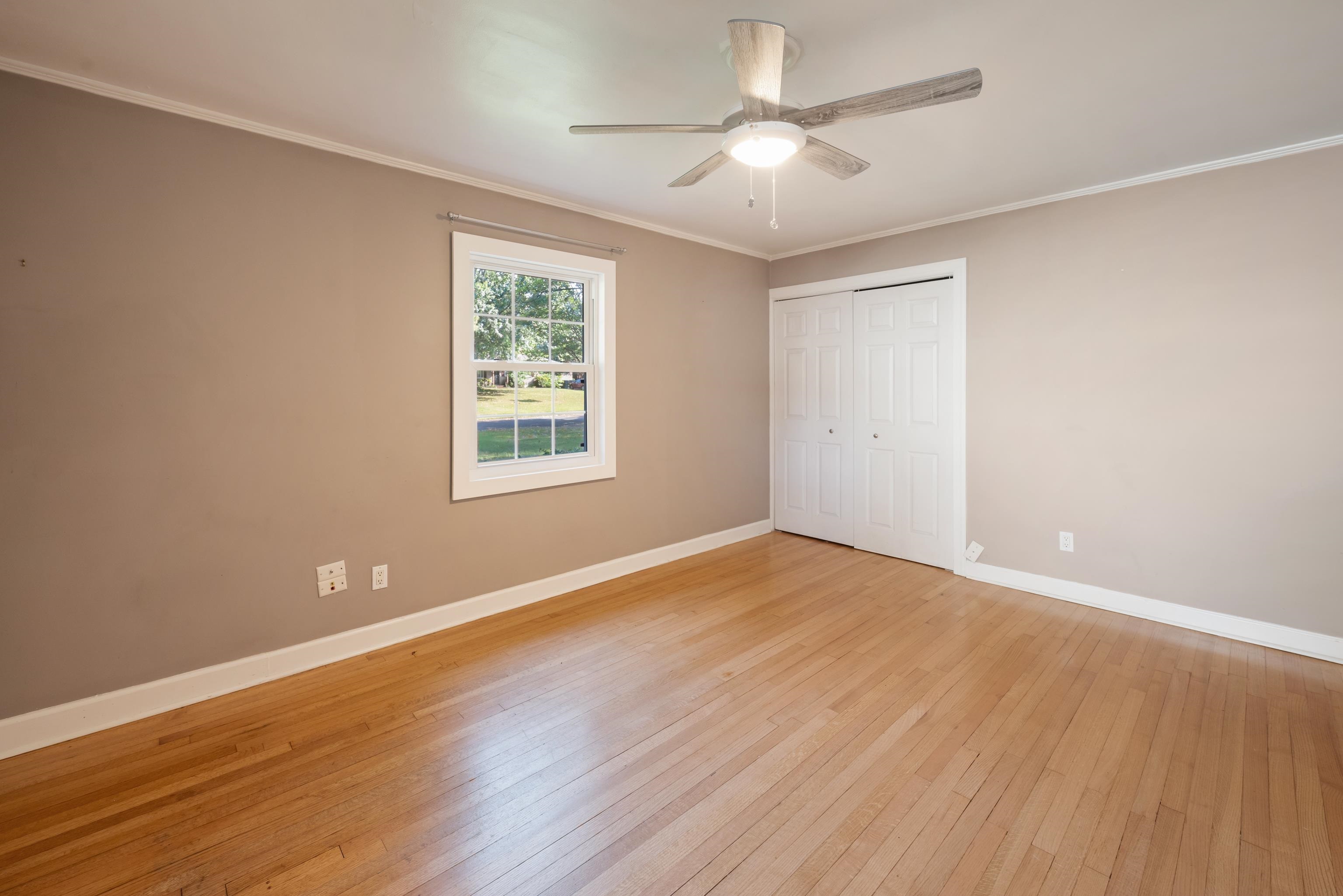 Empty bedroom with hardwood floors, a window, double closet doors, and a ceiling fan.