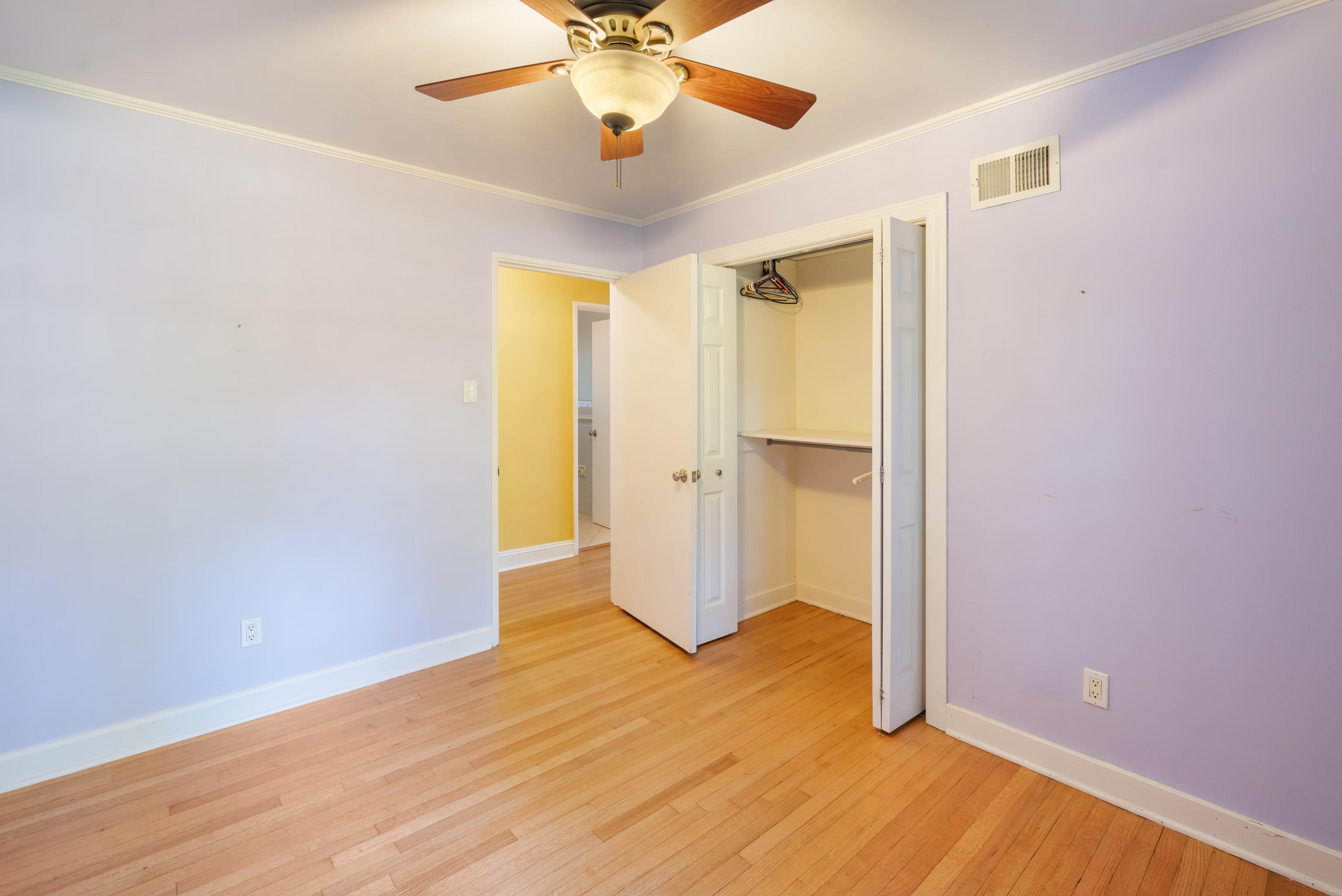 Empty bedroom with lavender walls, hardwood floors, ceiling fan, and open closet.