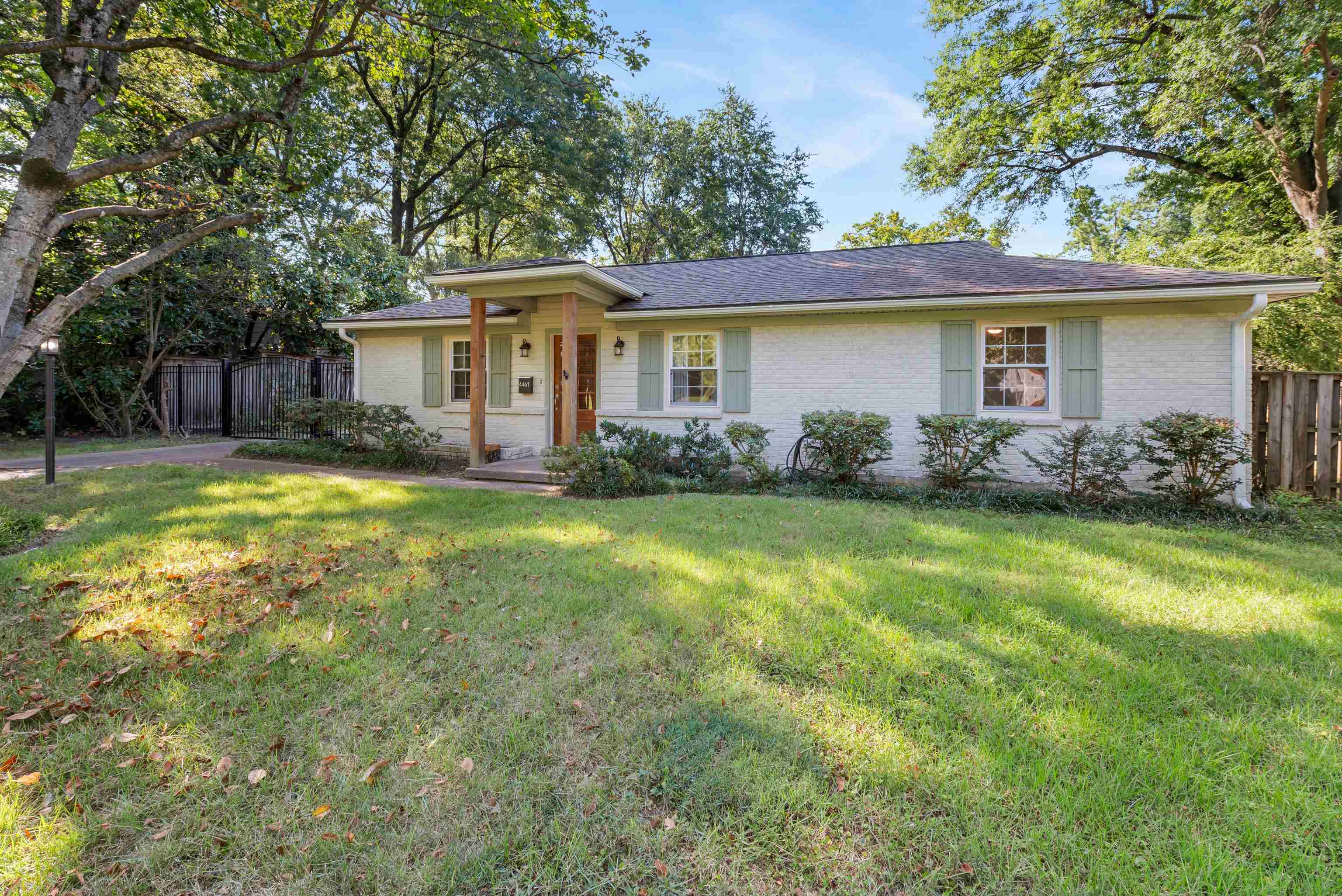 Single-story white-brick home with pale green shutters, a covered entry, and a broad lawn beneath mature trees.