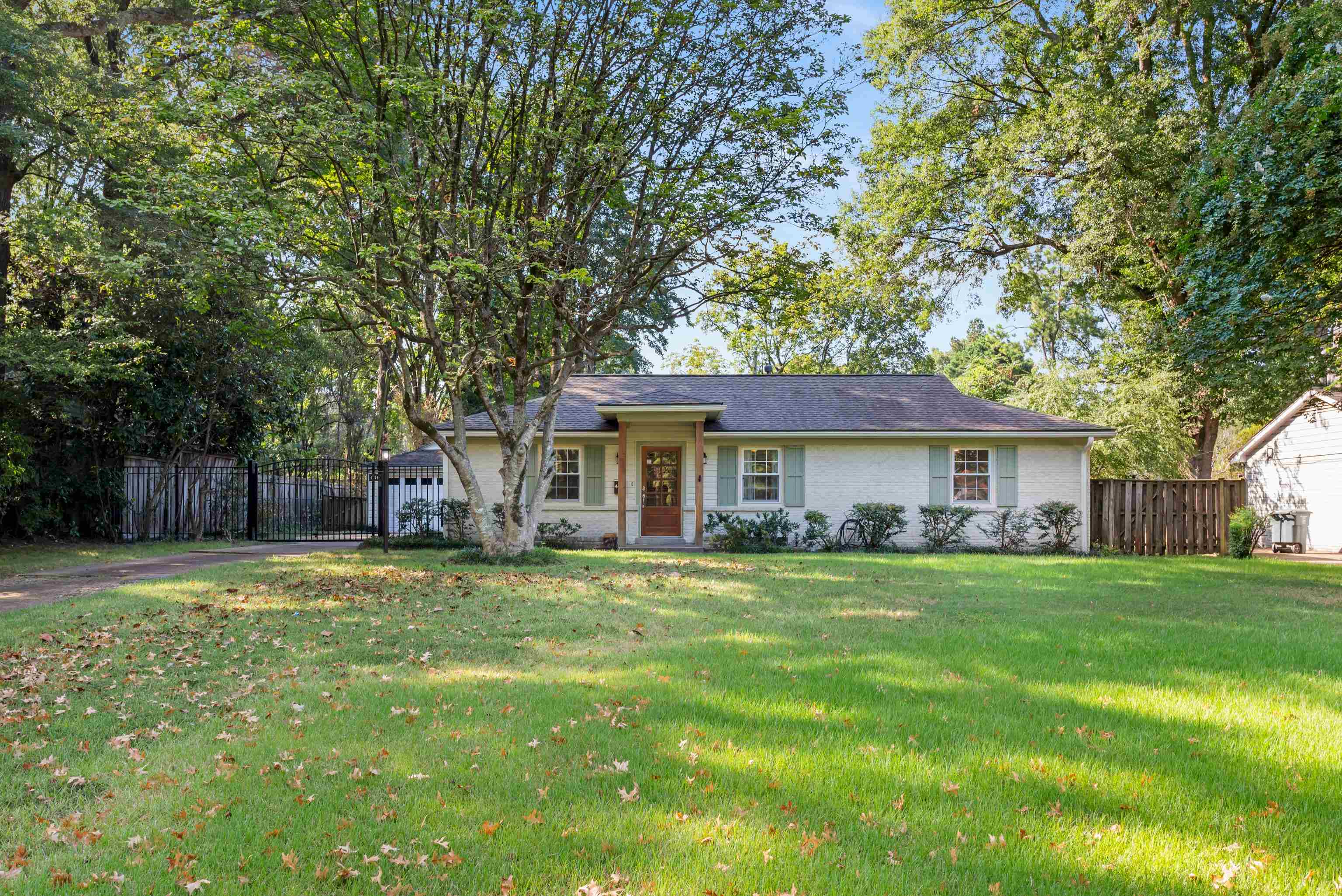 Single-story white brick house with pale green shutters, a wood entry door, and a shaded front lawn.