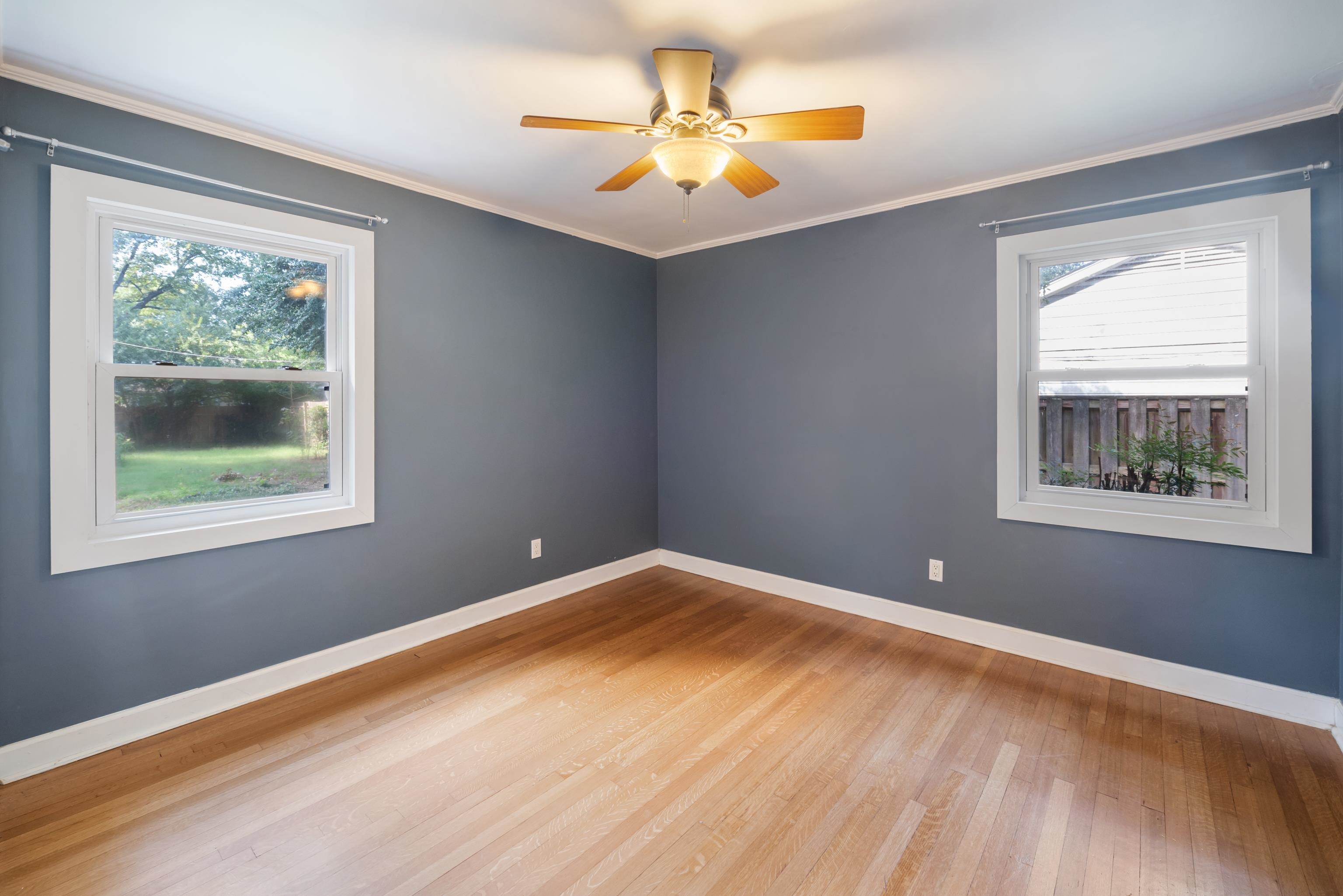 Vacant room with blue walls, hardwood floors, two windows, and a lit ceiling fan.
