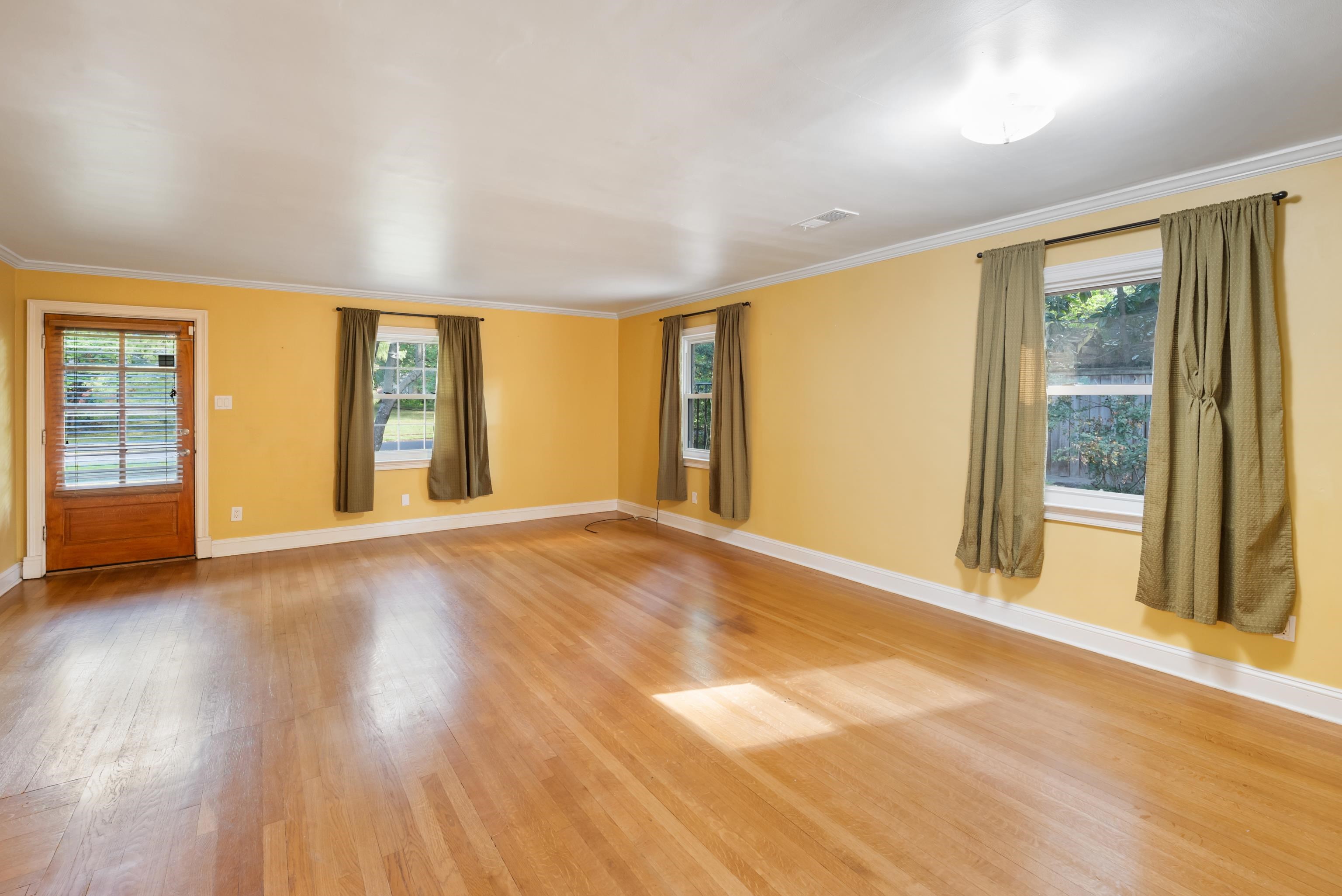 Empty room with yellow walls, hardwood floors, curtained windows, and a wooden door with blinds.
