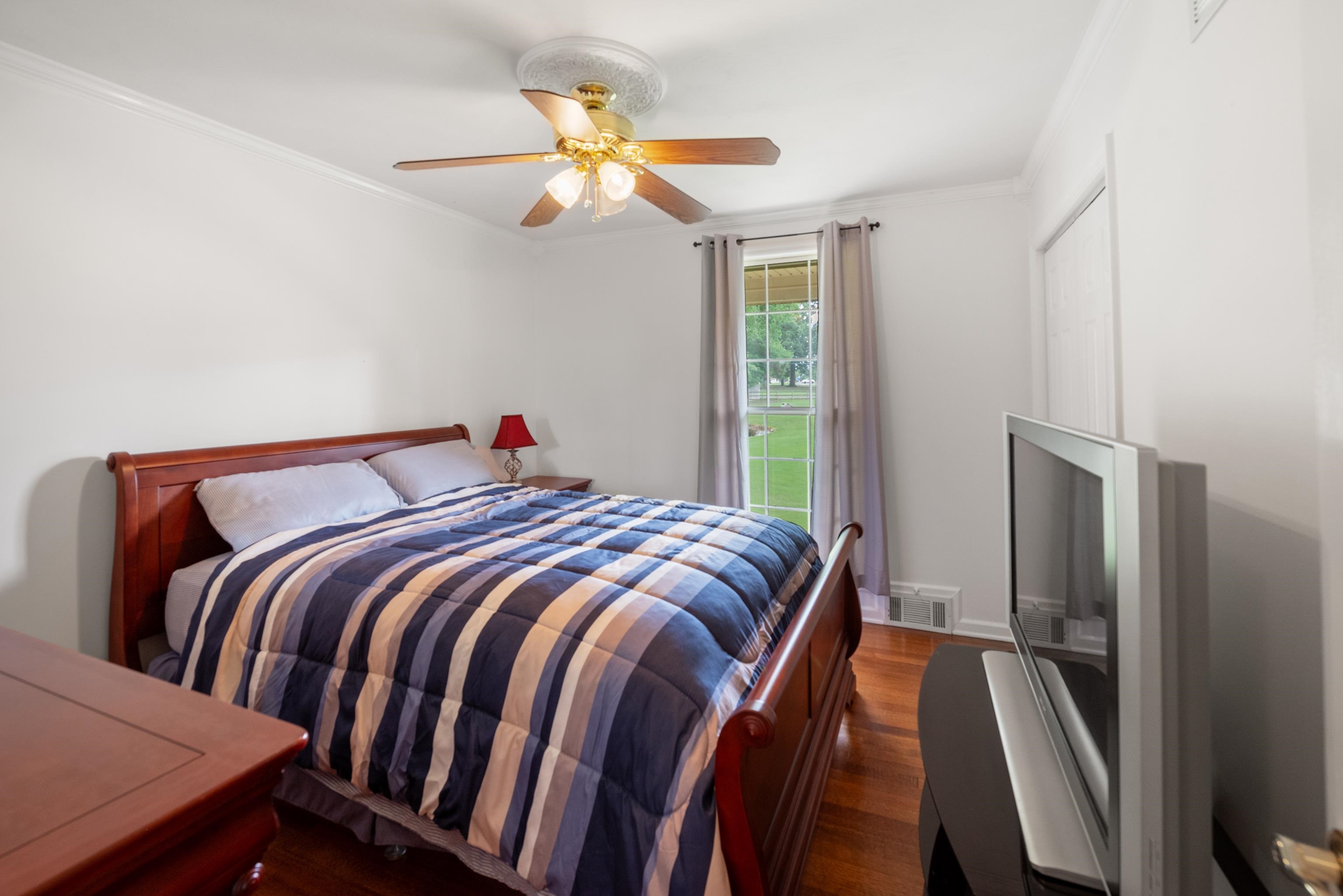 Bedroom with a wooden sleigh bed, striped quilt, hardwood floors, and a tall window overlooking a lawn.