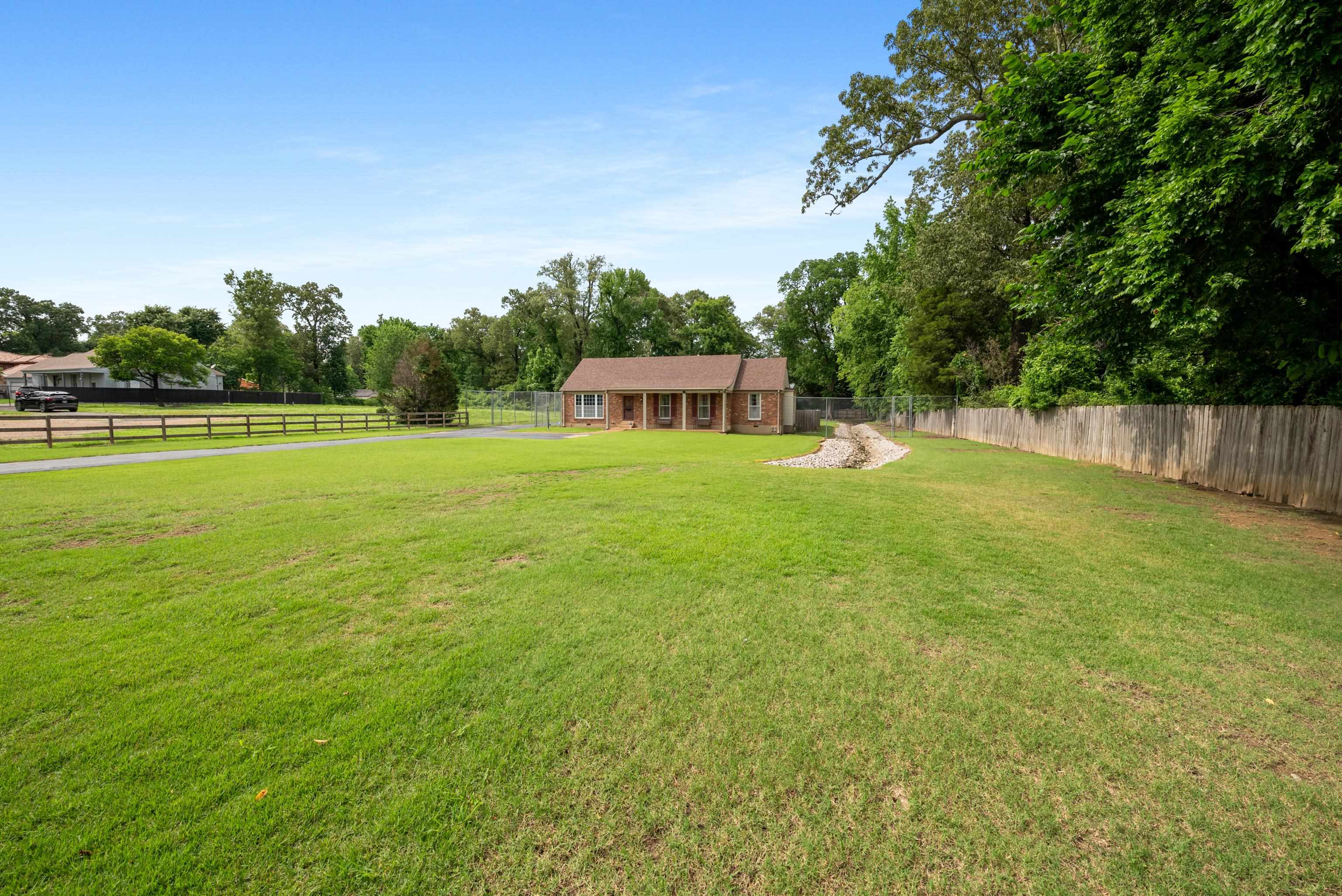 Brick house beyond a broad grassy yard, with mature trees and fencing along the property.