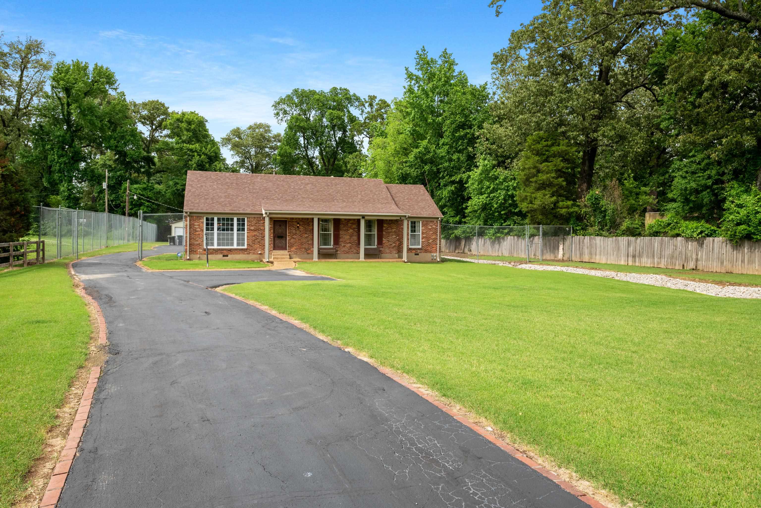Single-story brick home with a long curved driveway, fenced lawn, and mature trees.