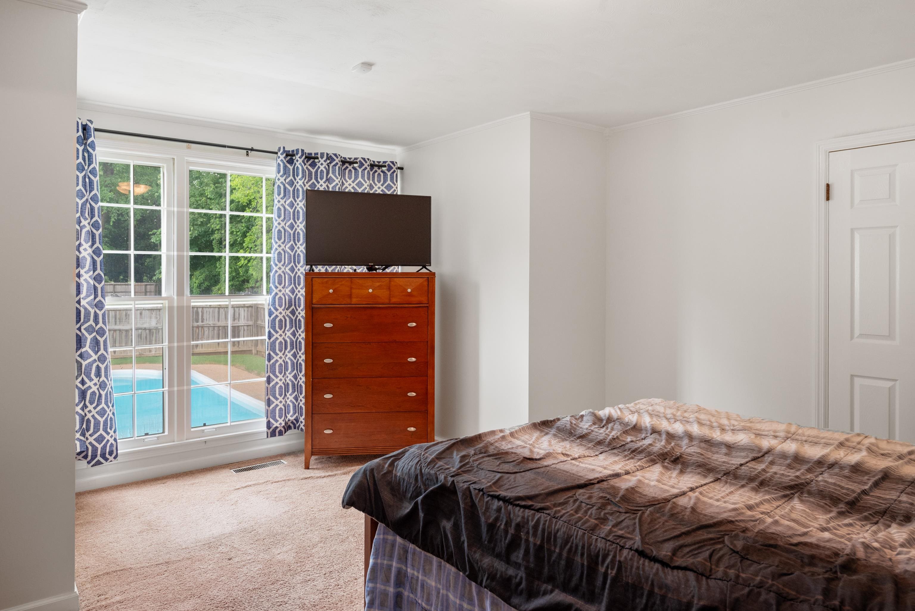 Bedroom with a wooden dresser and television beside large windows overlooking a backyard pool.