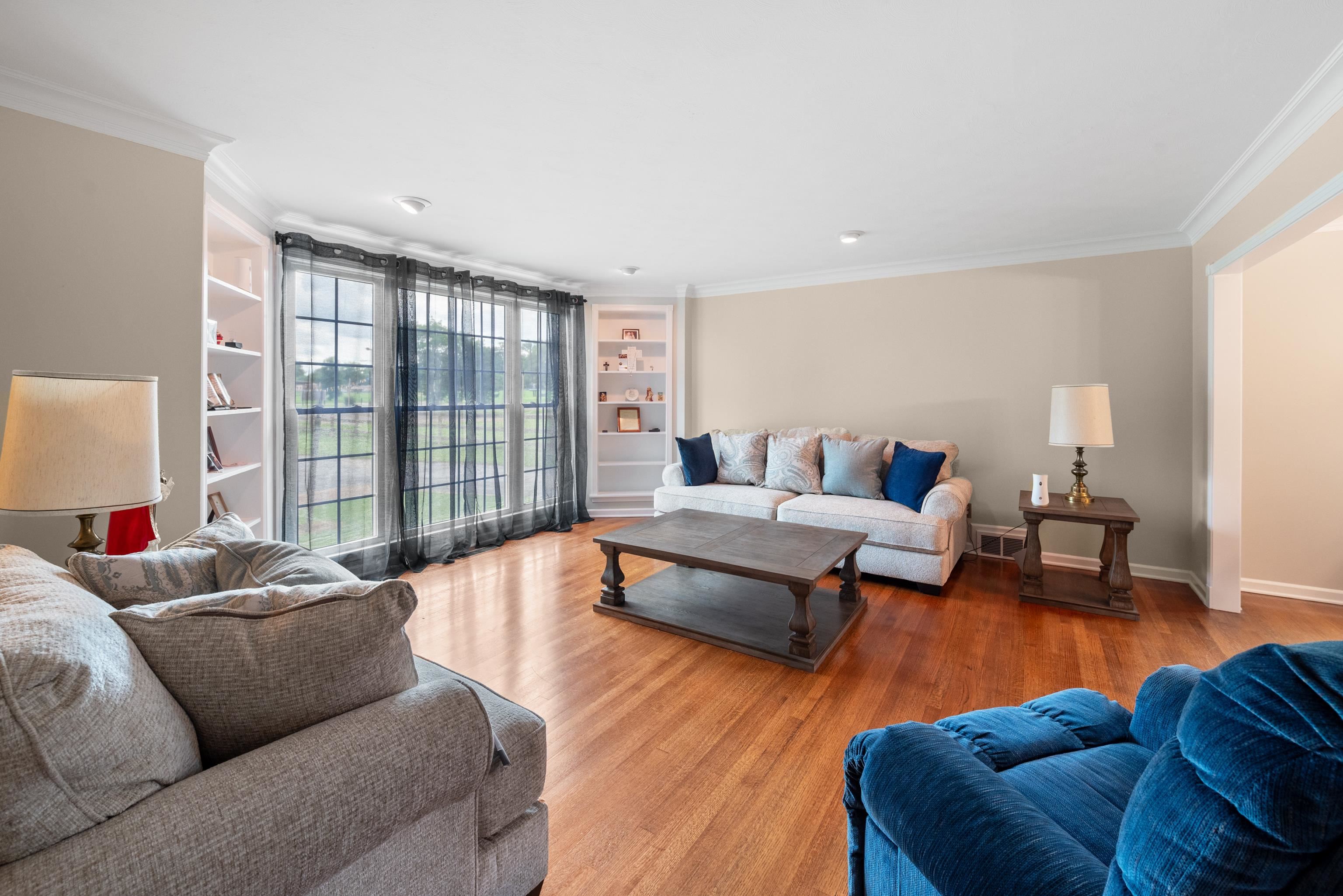 Living room with hardwood floors, large curtained windows, built-in shelves, and sofas around a coffee table.