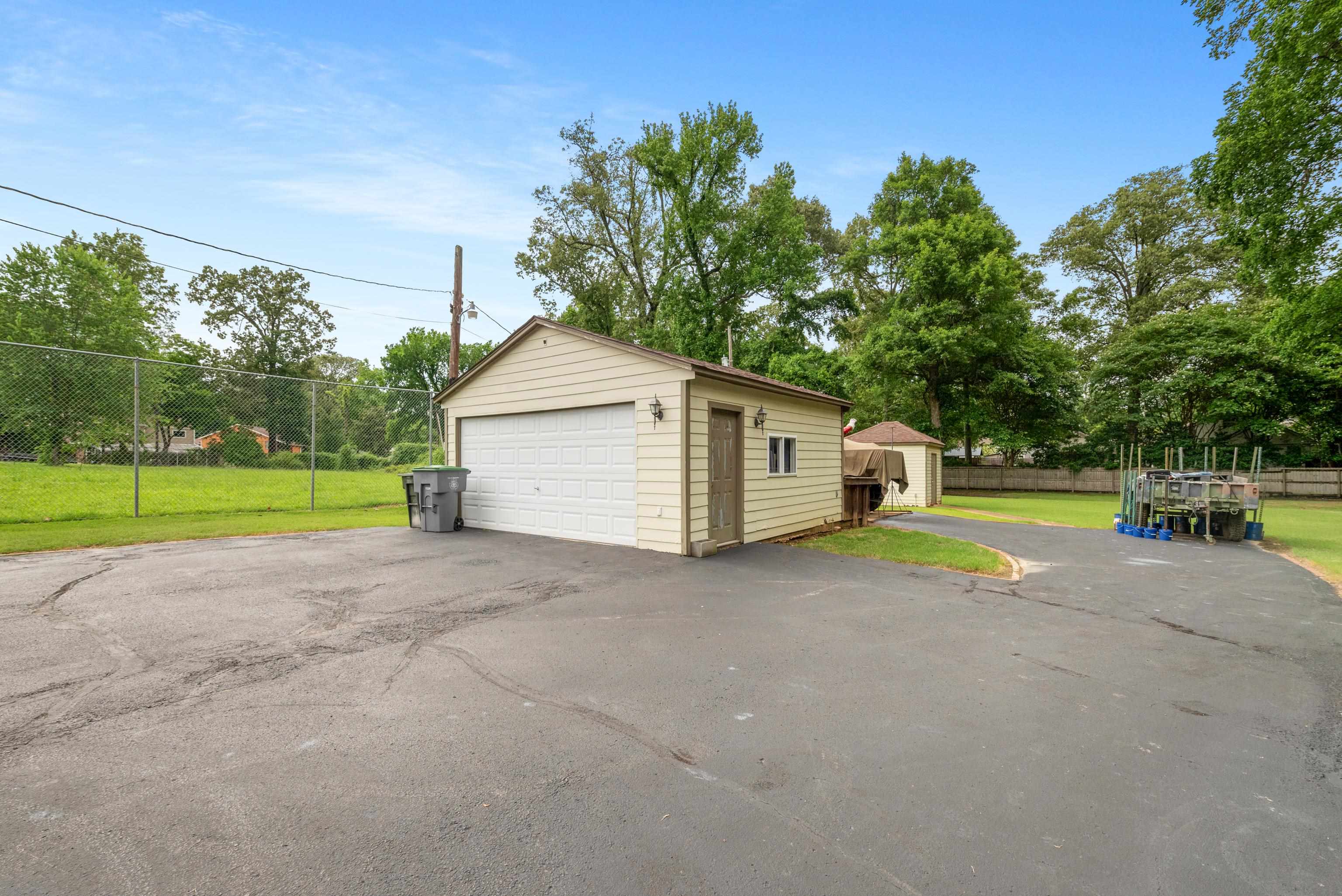 Detached beige garage with a white double door beside a broad asphalt parking area and fenced yard.