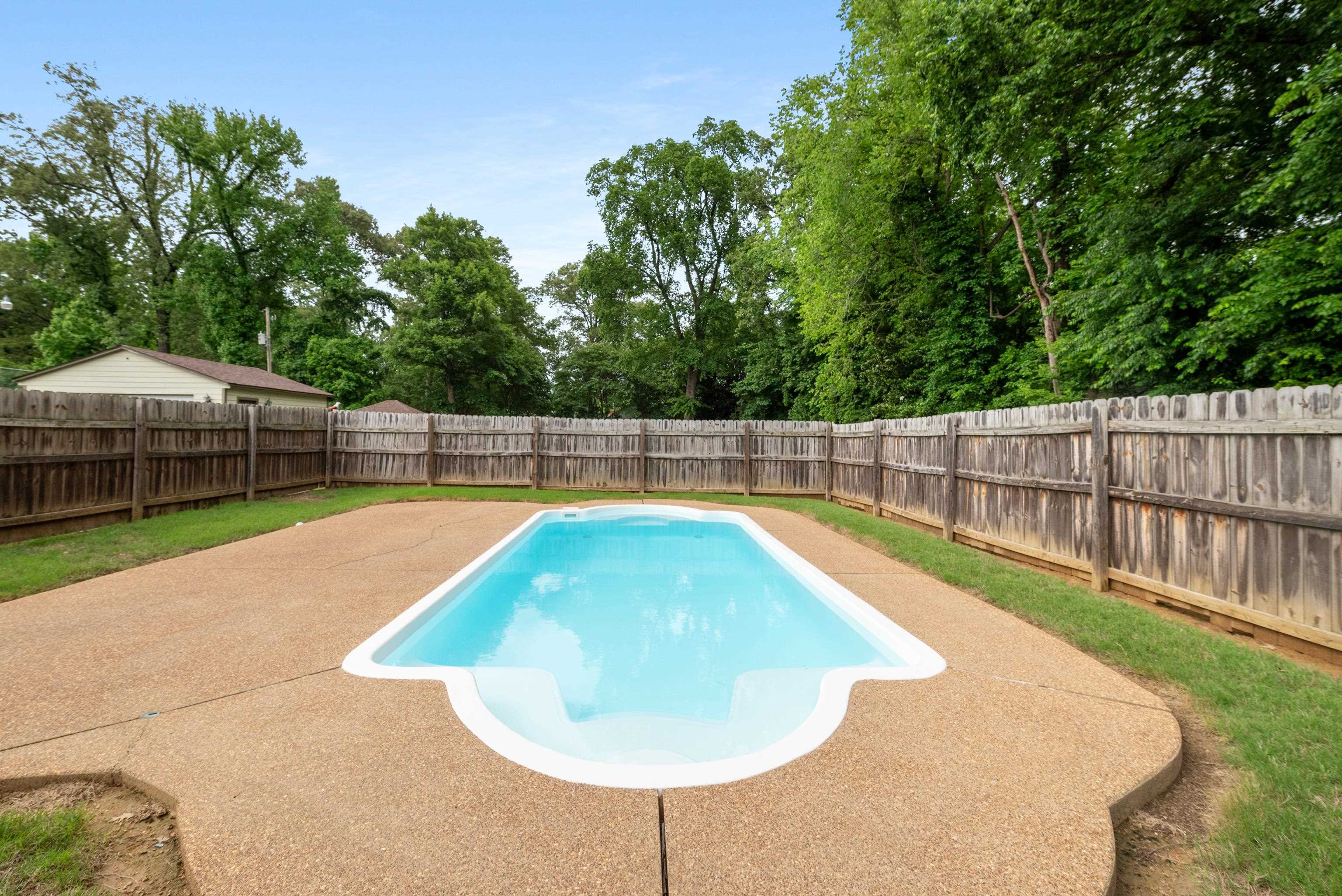 Backyard swimming pool with a broad concrete deck and wooden privacy fence beneath mature trees.