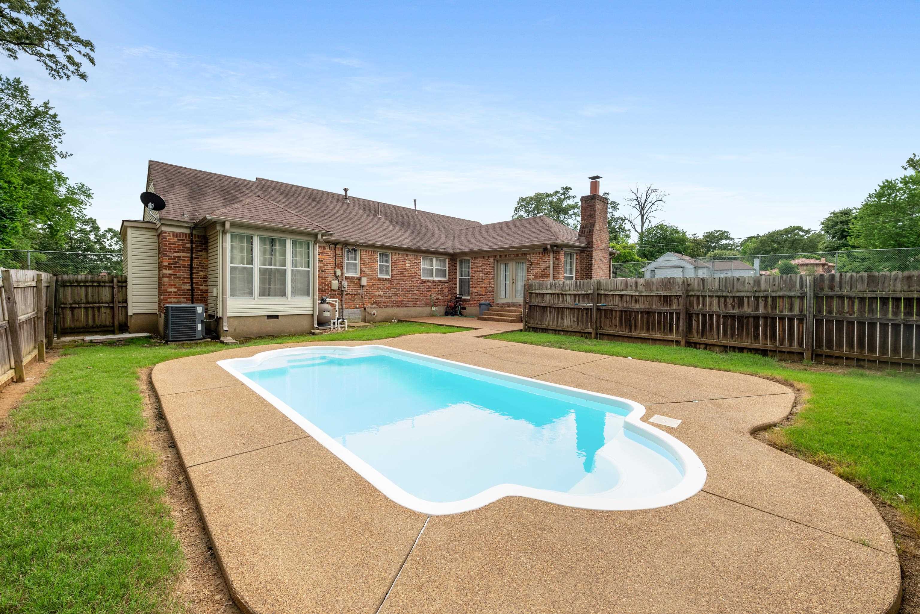 Backyard pool and concrete deck behind a brick house, enclosed by a wooden privacy fence.