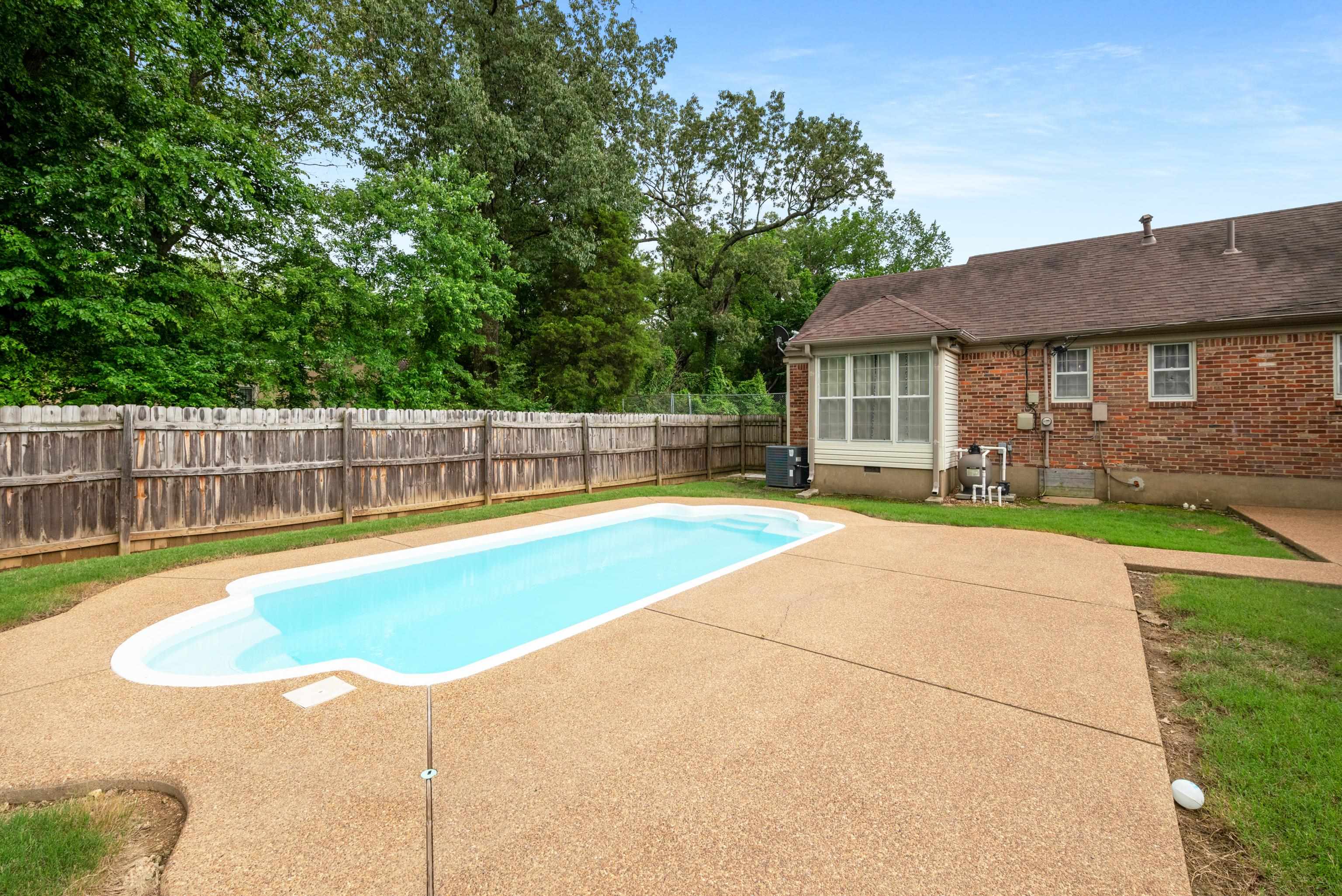 Backyard swimming pool beside a brick house, with a wide concrete patio and fenced yard.