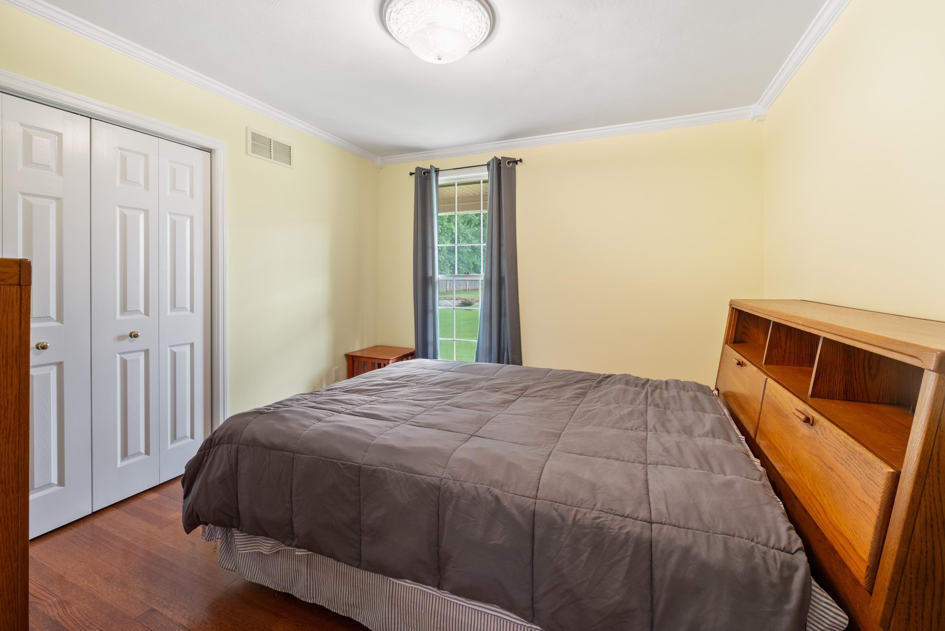 Furnished bedroom with a gray-quilted bed, white closet doors, and a narrow window overlooking the lawn.