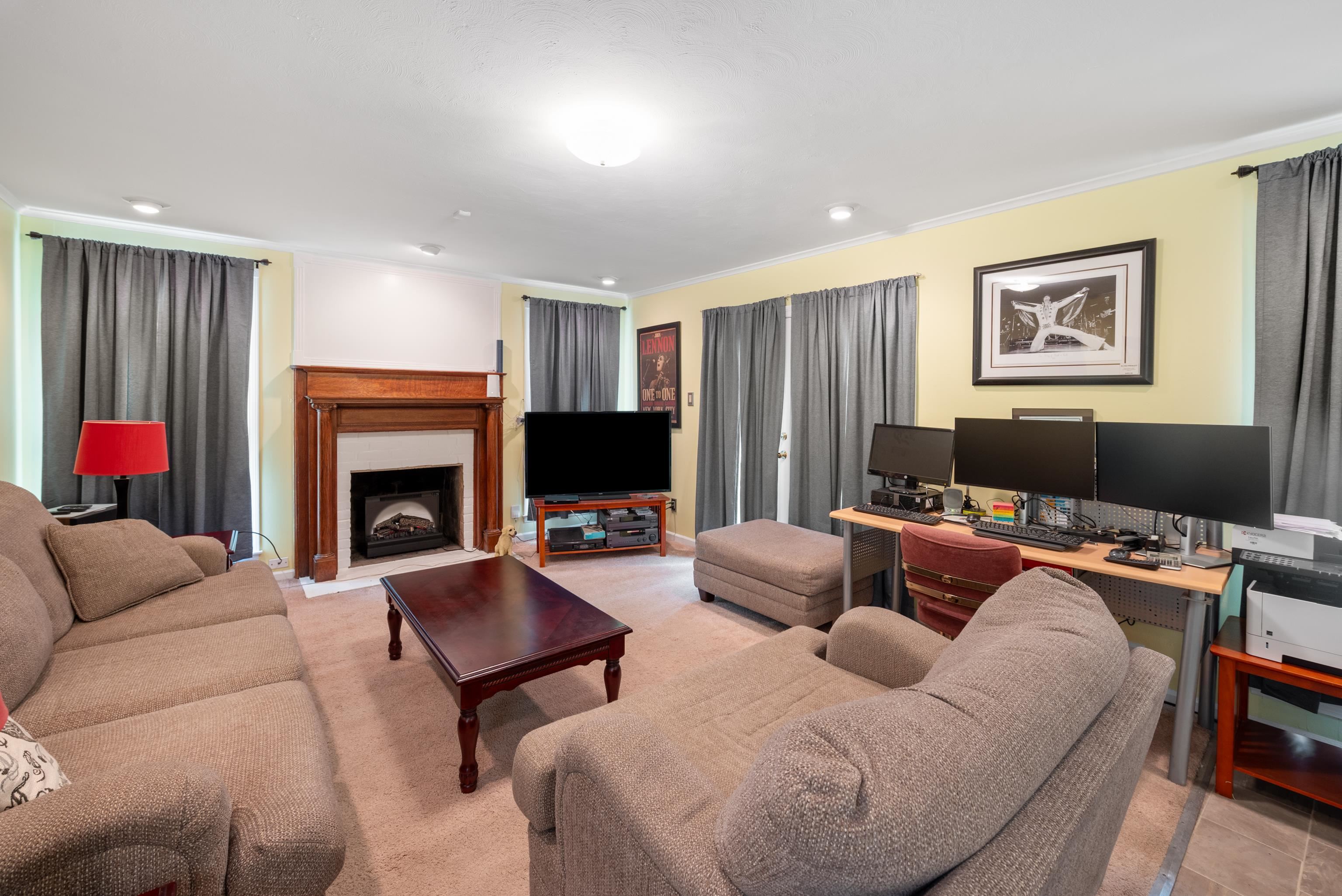 Living room with a wood fireplace, beige sofas, and a computer workstation with multiple monitors.
