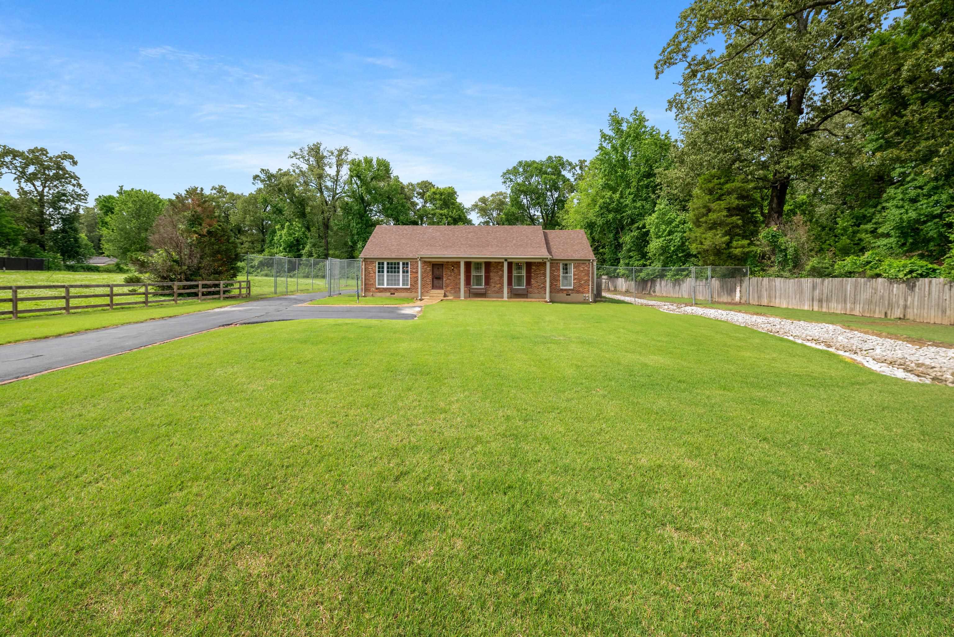 Single-story brick house beyond a broad lawn, with a curved driveway and fenced yard.