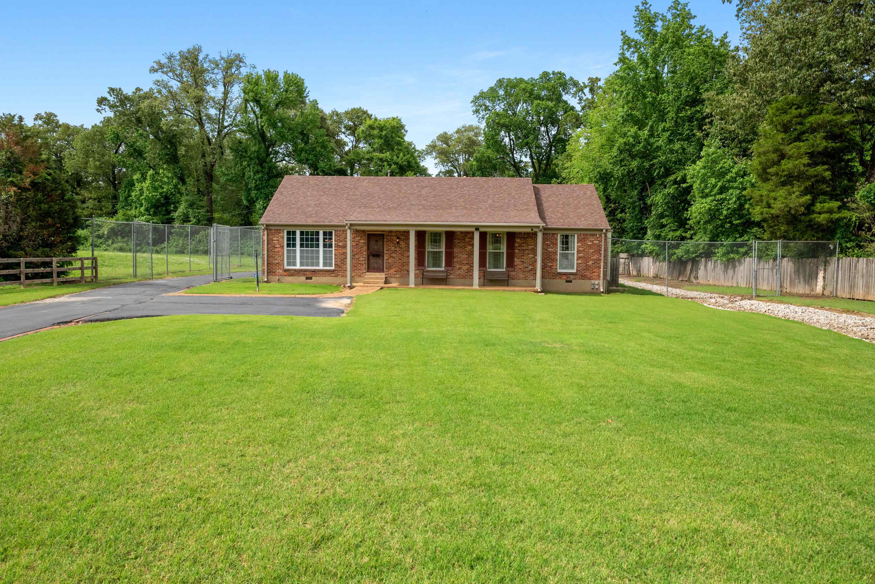 Single-story brick house with a covered porch, broad lawn, paved driveway, and fenced yard.
